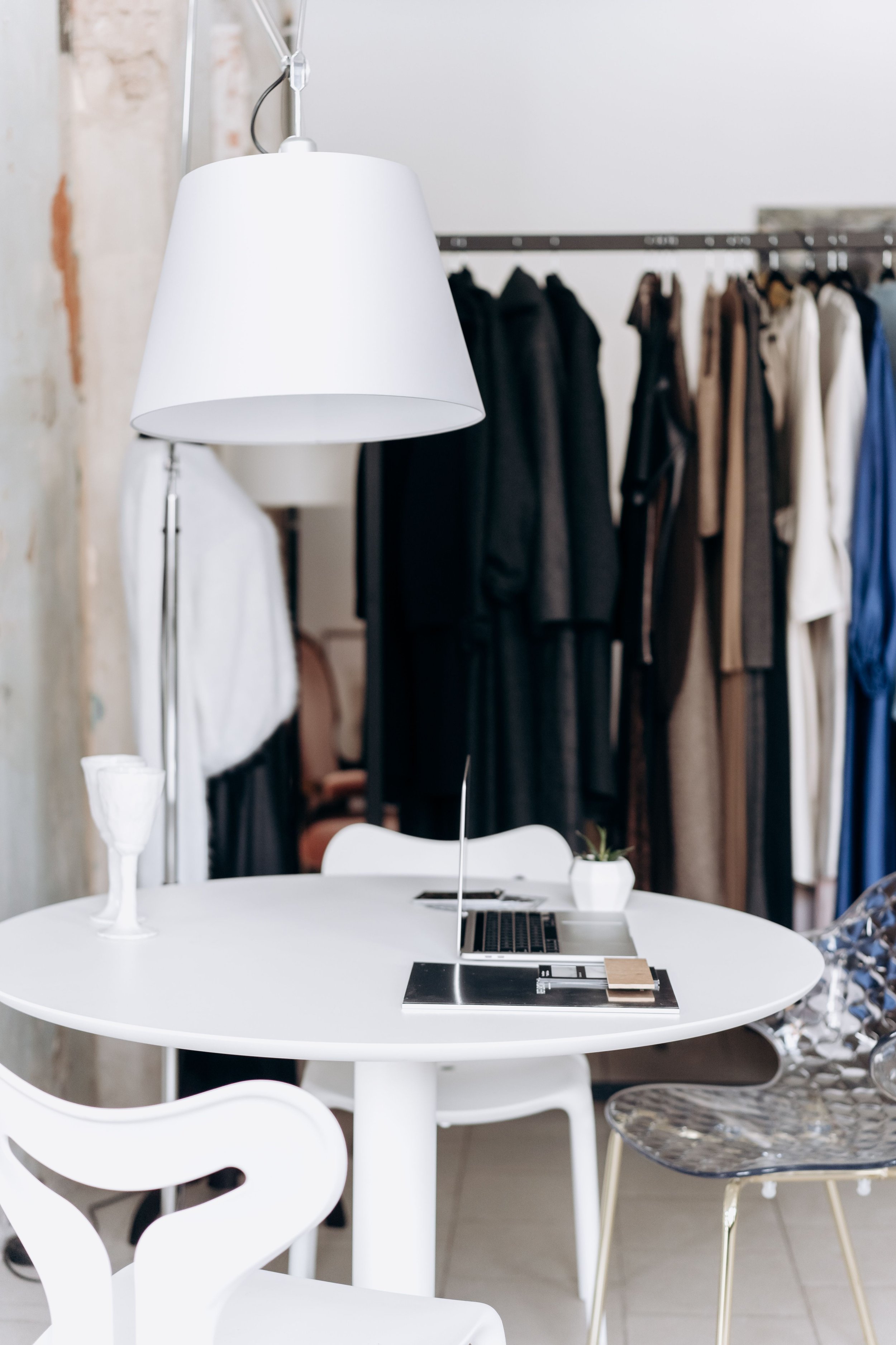 A modern workspace with a white round table, a laptop, a plant, and several chairs in a bright room. In the background, there are black and beige clothing items hanging on racks and a white wall.