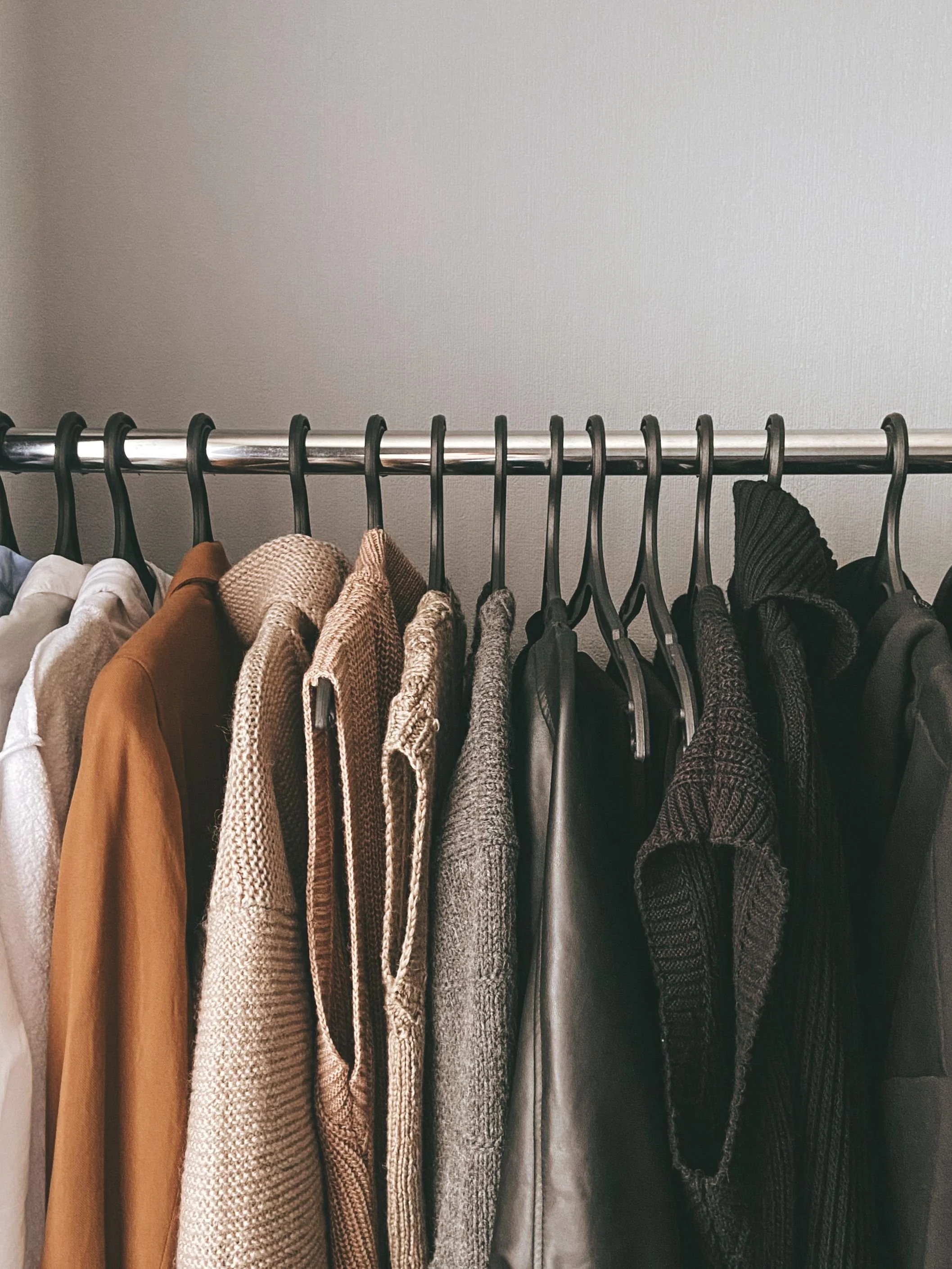 Clothing hanging on a metal rod in a closet, including sweaters, jackets, and a scarf, against a plain wall.