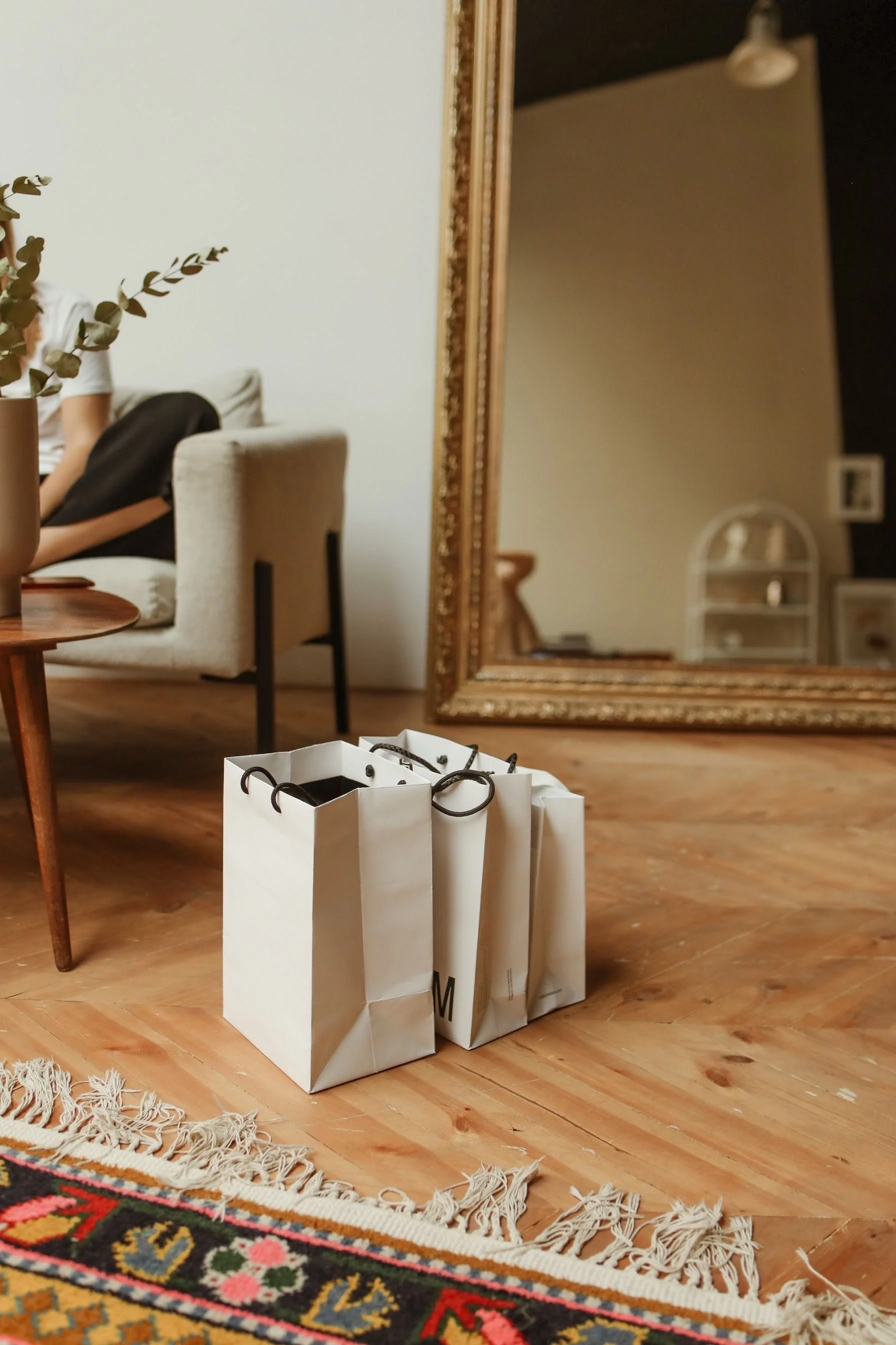 Three white shopping bags with black handles sitting on a wooden floor near a colorful fringed rug, with a large ornate mirror and a beige upholstered chair visible in the background.