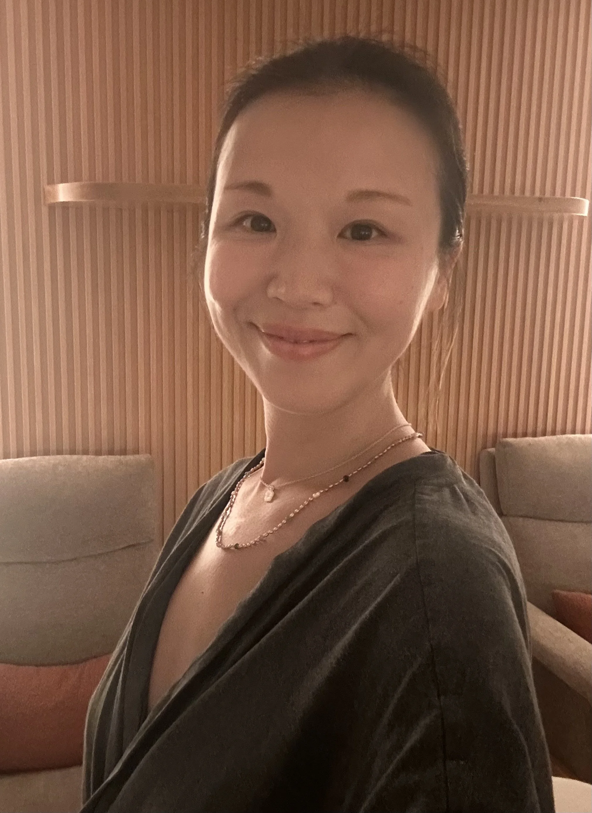 A young woman with dark hair tied back, smiling, wearing a black top and layered necklaces, sitting in a room with wooden paneling on the wall and beige couches.