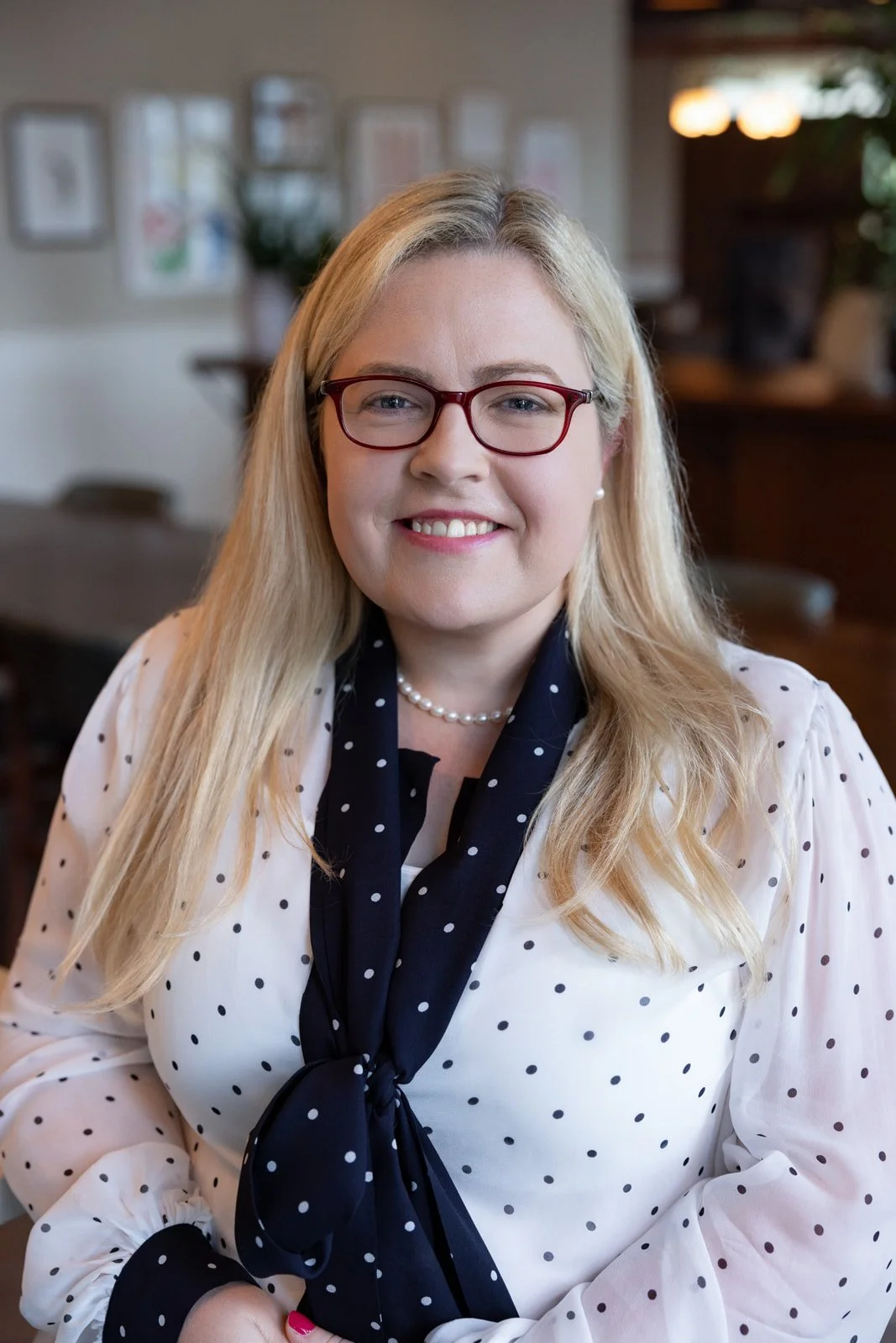 A woman with blonde hair, red glasses, and a pearl necklace, smiling while sitting in a warmly-lit room with framed pictures on the wall.