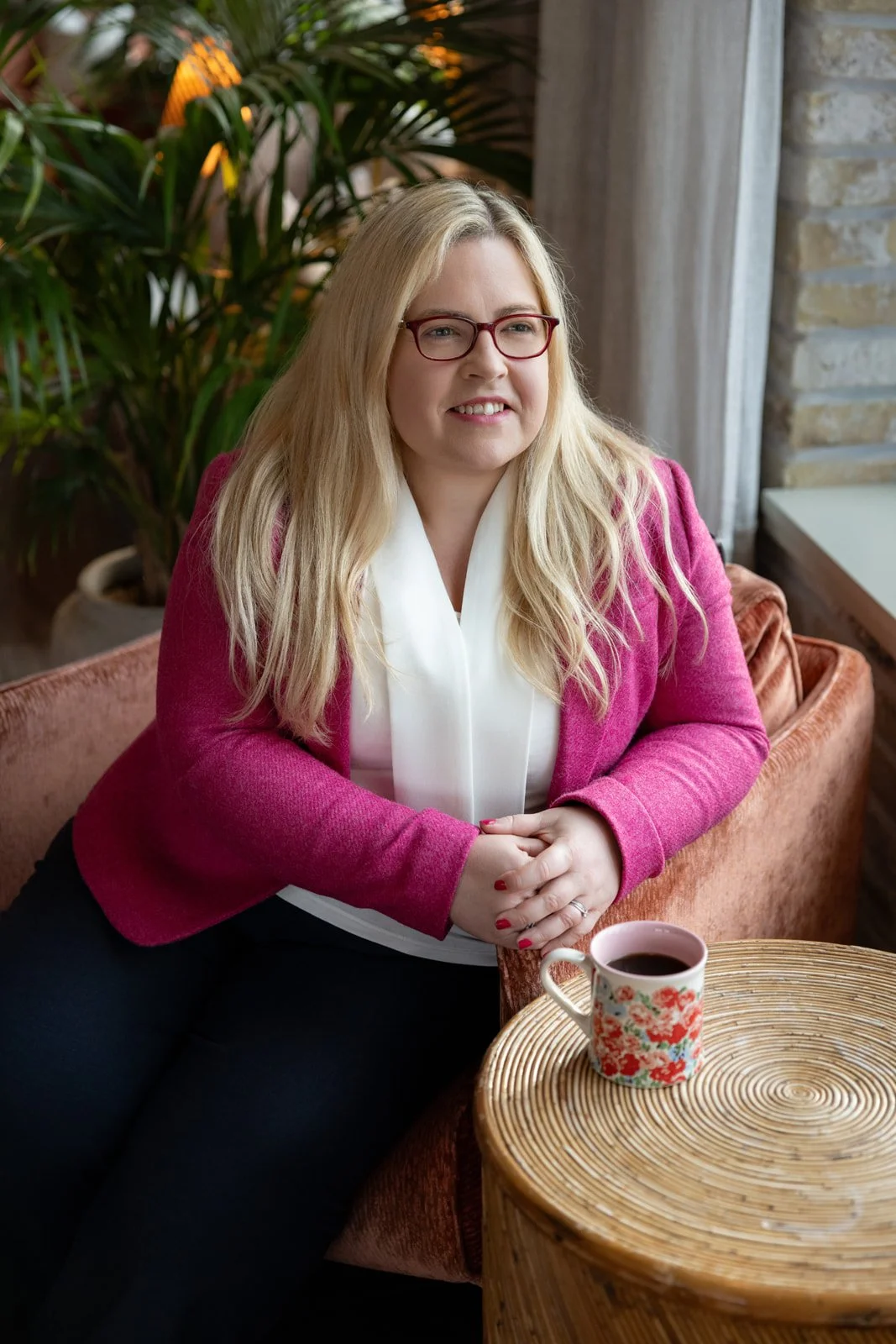 A woman with blonde hair, wearing glasses, a white top, and a pink jacket, sitting on a pinkish-orange couch with a cup of coffee on a round wooden table in front of her, in a cozy cafe setting with plants and brick wall in the background.