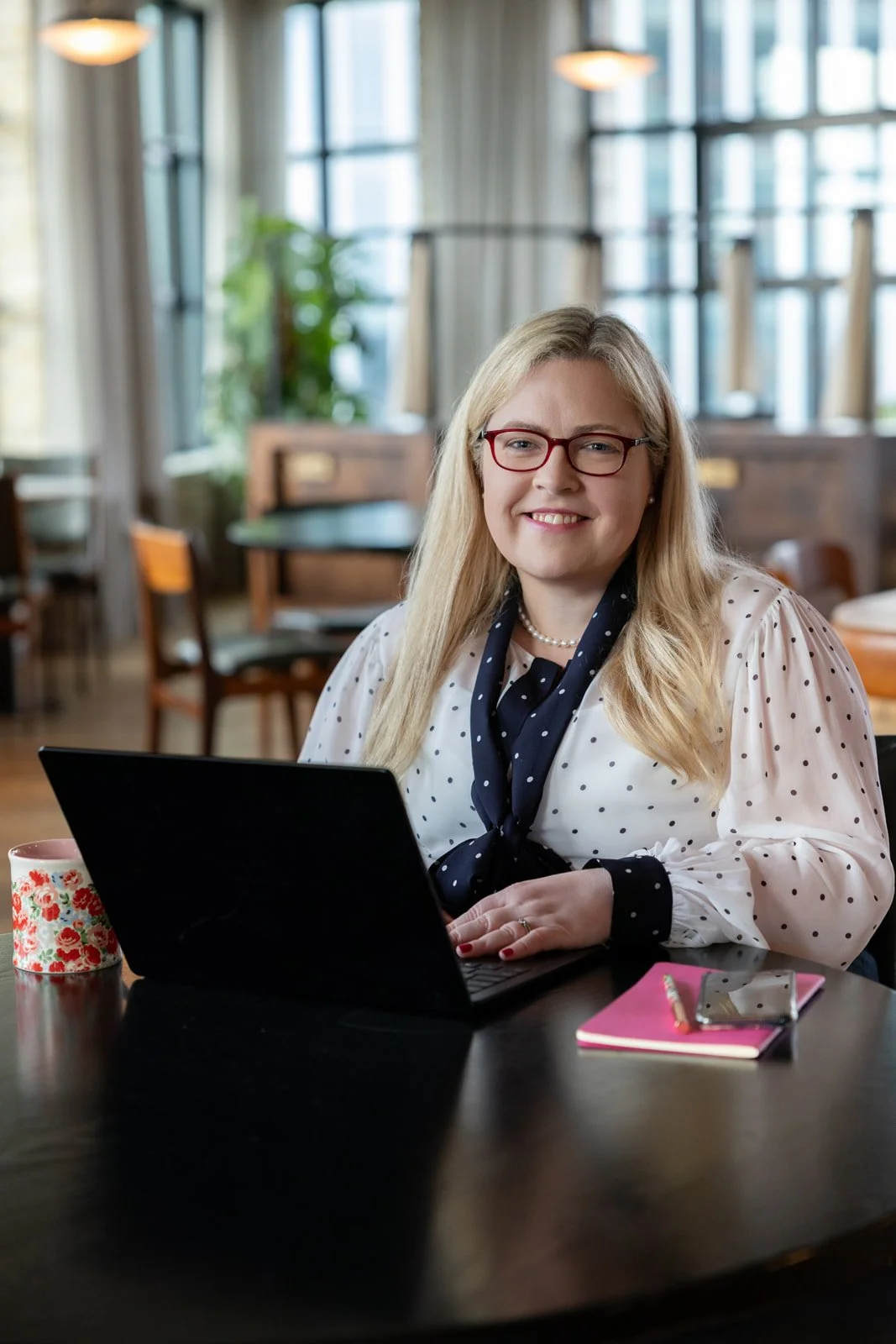 A smiling woman with blonde hair, wearing glasses and a white blouse with black polka dots, sitting at a table with a laptop, in a bright, cozy cafe.