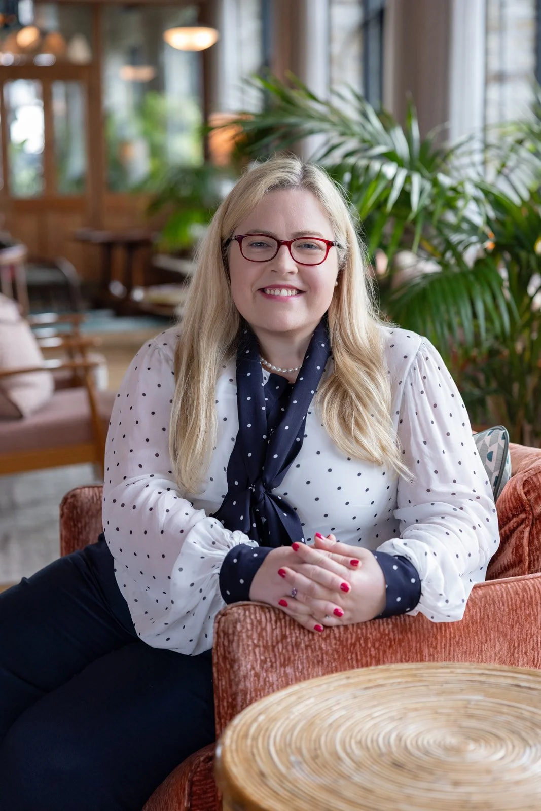 A smiling woman with blonde hair and red glasses sitting on a pink upholstered armchair in a cozy indoor setting, surrounded by greenery and wooden furniture.