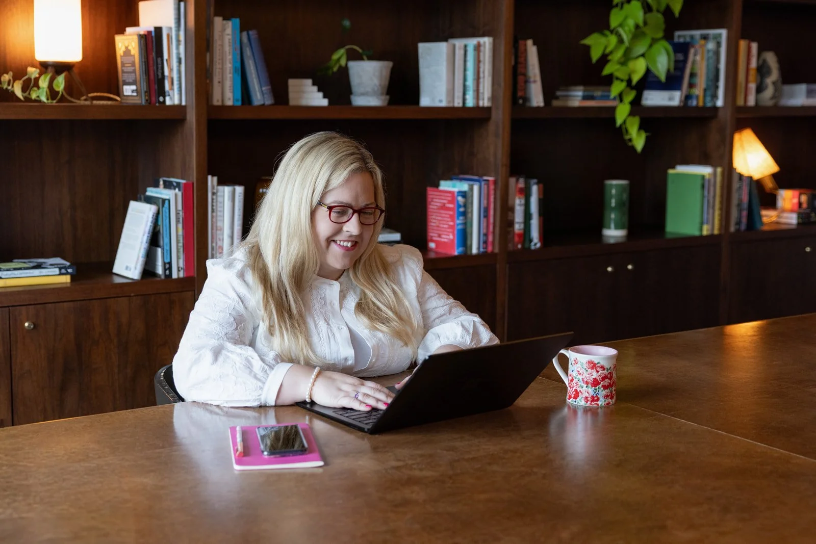 A woman with long blonde hair, glasses, and a white blouse sitting at a wooden table, smiling while using a laptop. There is a pink notebook with a phone on top and a floral mug on the table. Behind her, a bookshelf filled with books and plants is visible.