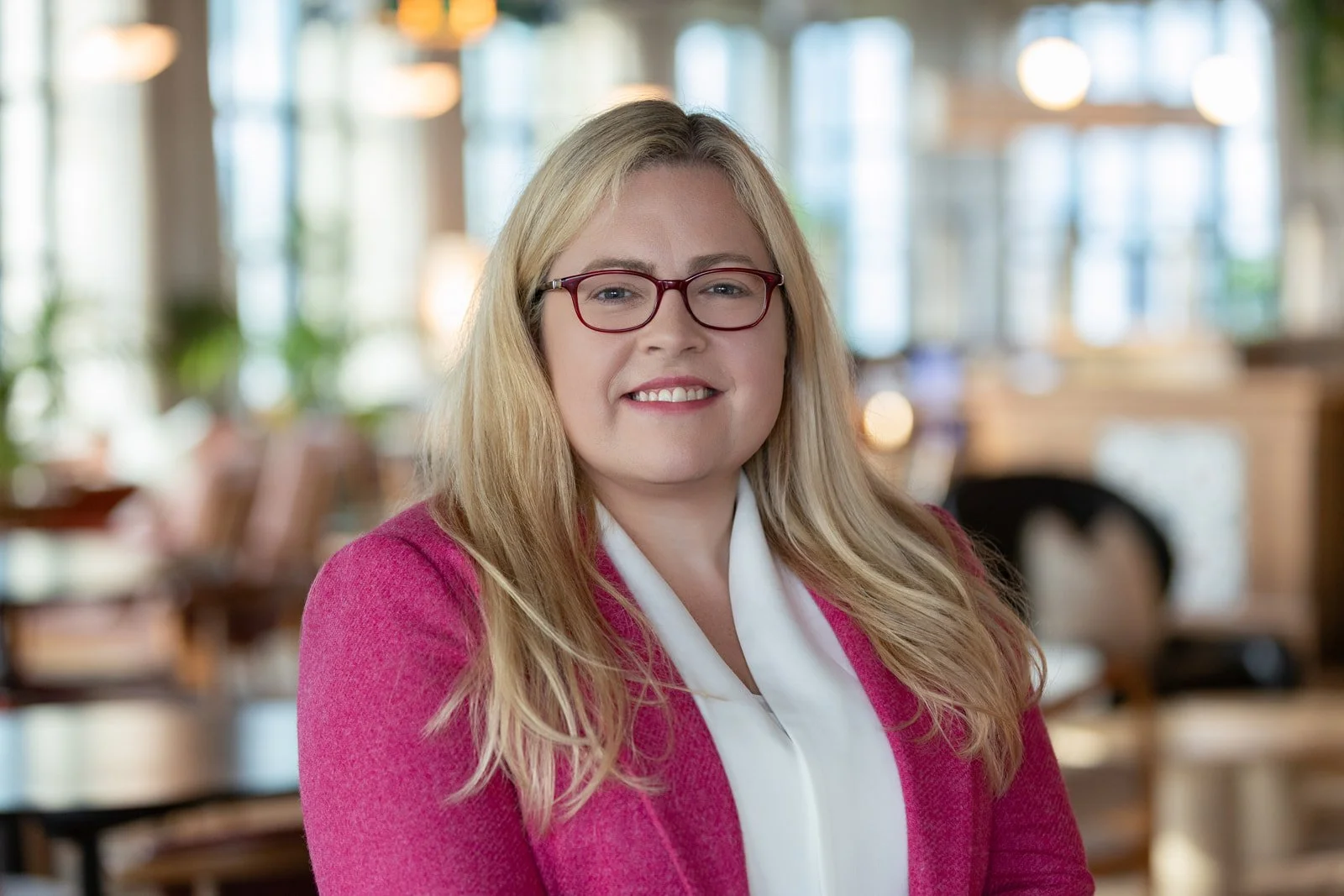 A woman with blonde hair and glasses smiling at the camera, wearing a pink blazer and white blouse, in a brightly lit cafe or restaurant.