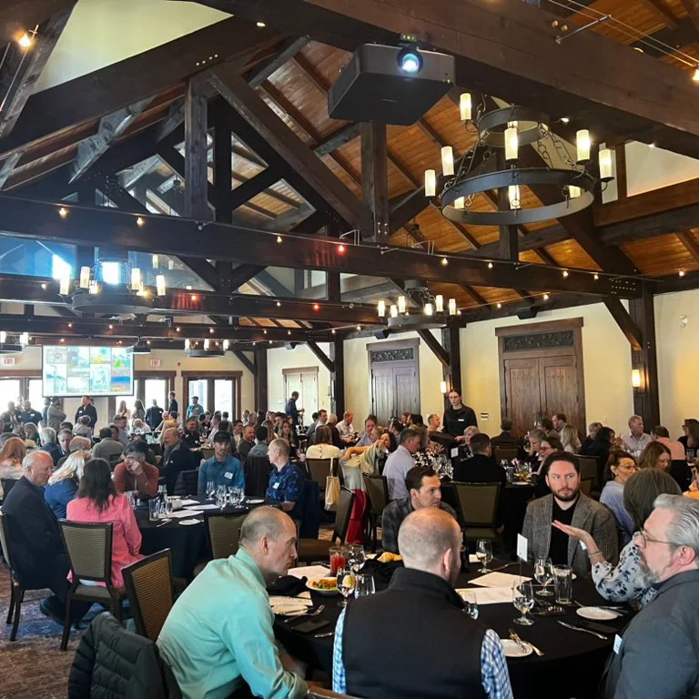 Crowded restaurant dining room with guests seated at round tables beneath rustic timber beams and chandeliers
