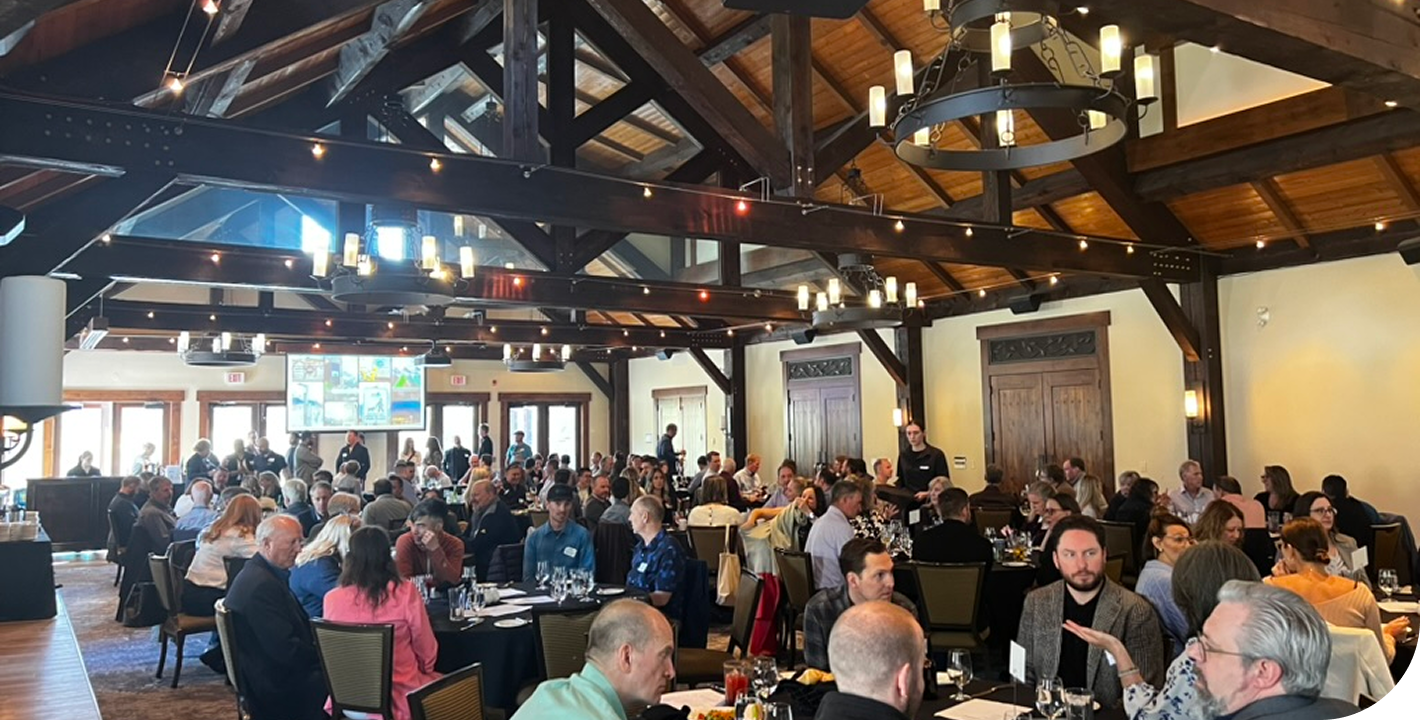 A large indoor banquet hall filled with people sitting at round tables, attending an event or conference, with a high wooden ceiling and large windows allowing natural light.
