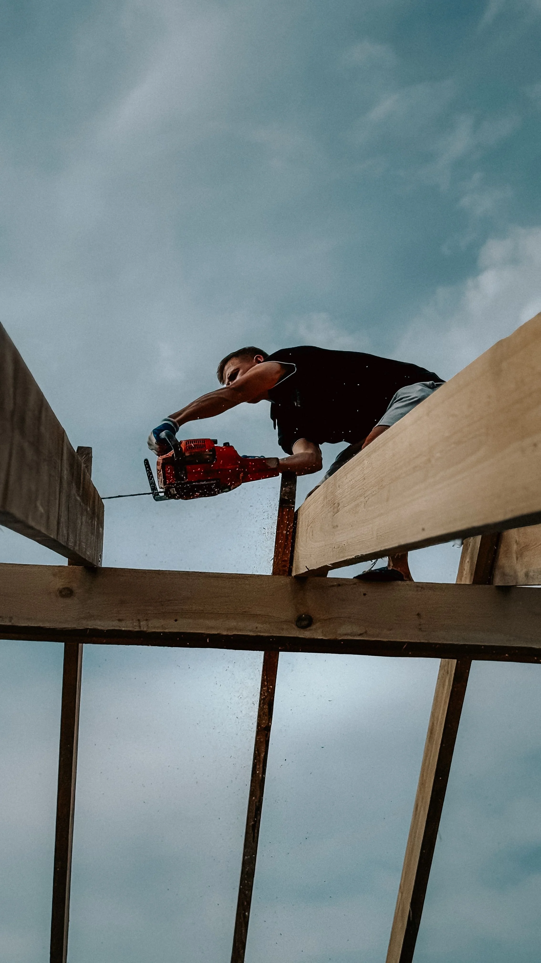 A man using a chainsaw to cut a wooden beam on a construction site outdoors under a cloudy sky.
