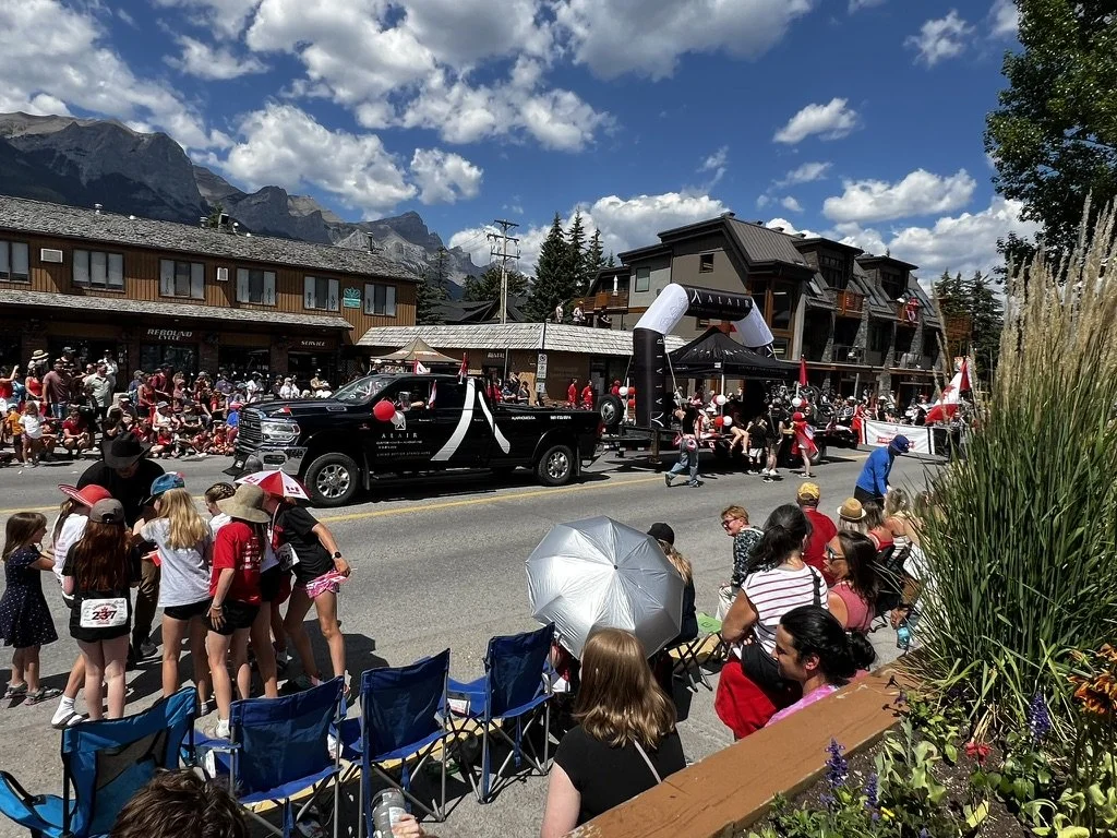 A parade on a street with spectators sitting and standing, a black truck with decorations, people in red shirts, and a building with mountain scenery in the background.