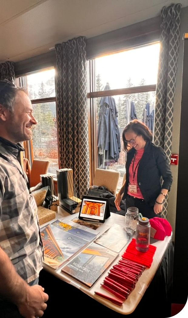 Two people smiling and talking at a conference table with brochures, a tablet, and red pens, inside a room with large windows and outdoor umbrellas.