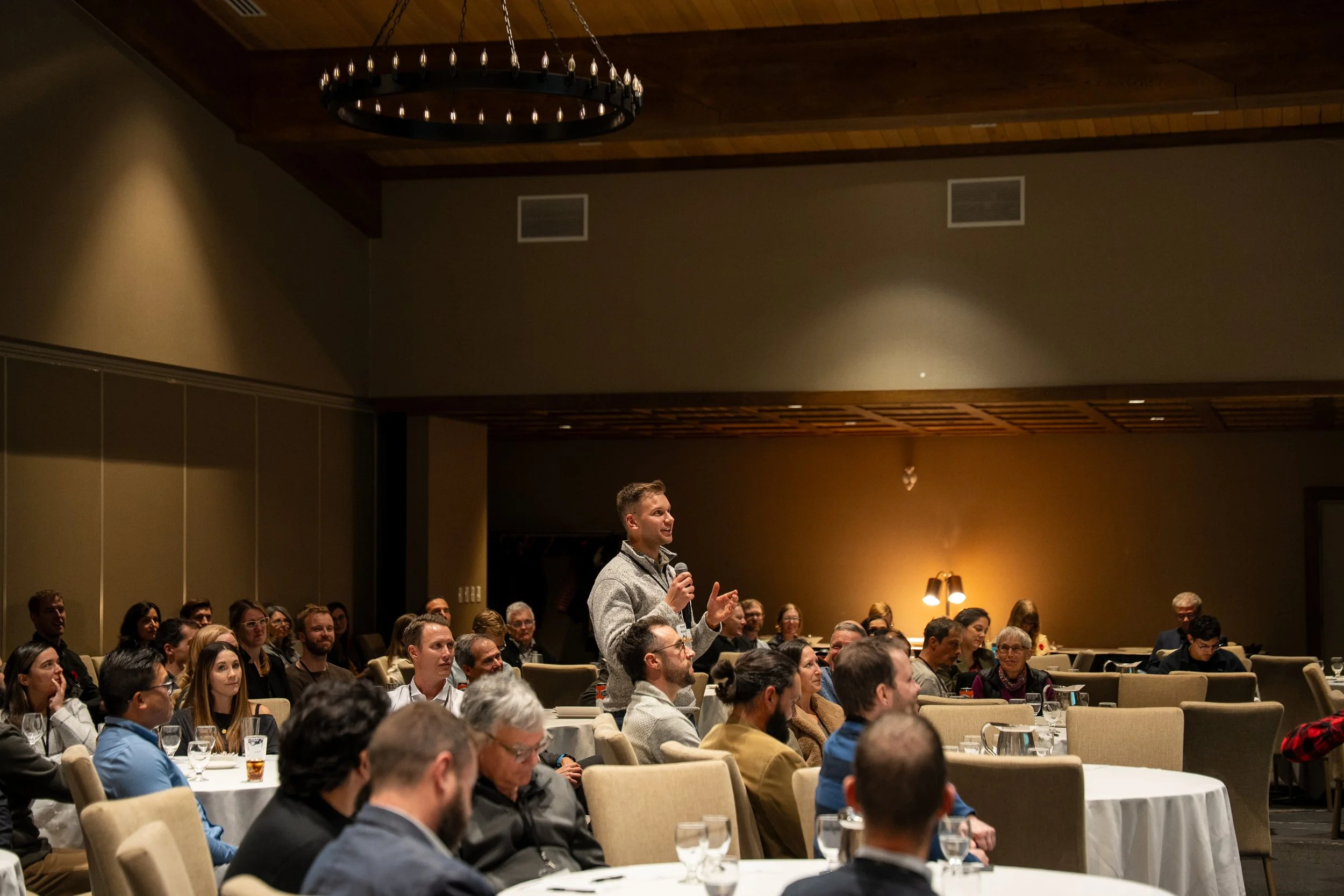 A man standing and speaking into a microphone during a conference or seminar, with an audience seated and listening attentively in a large, dimly lit room with a chandelier hanging from the ceiling.
