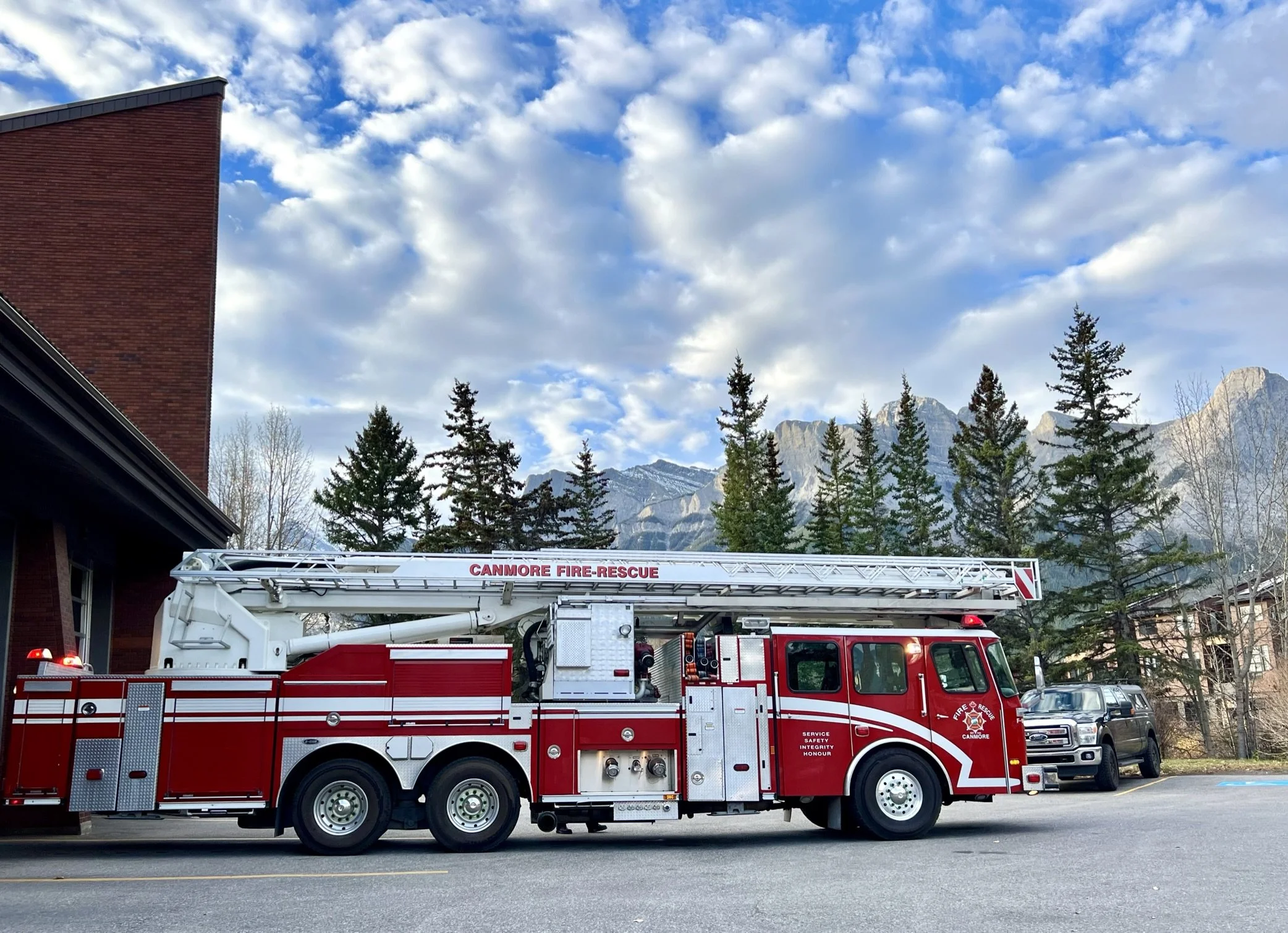 A red and white fire truck from the Canmore Fire-Rescue department parked on a street, with a backdrop of trees, mountains, and a partly cloudy sky.