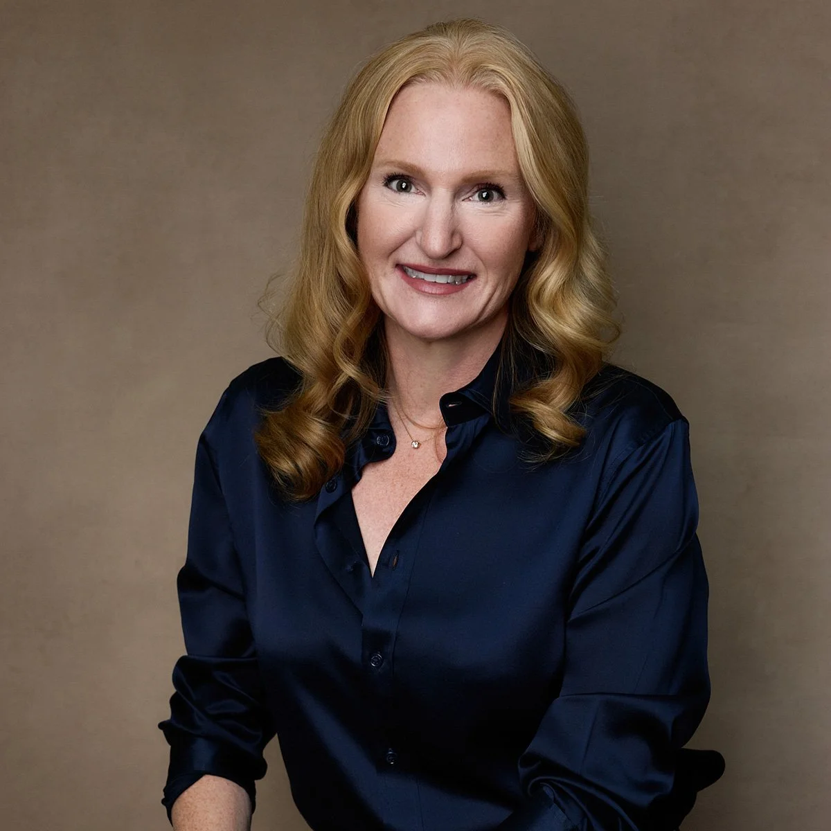 Professional portrait of a smiling blonde woman wearing a navy blouse against a brown background