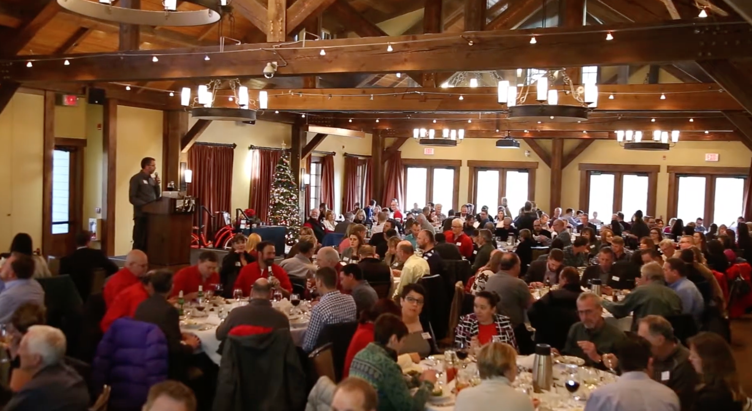 Large indoor gathering with people seated at tables, decorated for Christmas, with a Christmas tree, and a speaker at a podium.