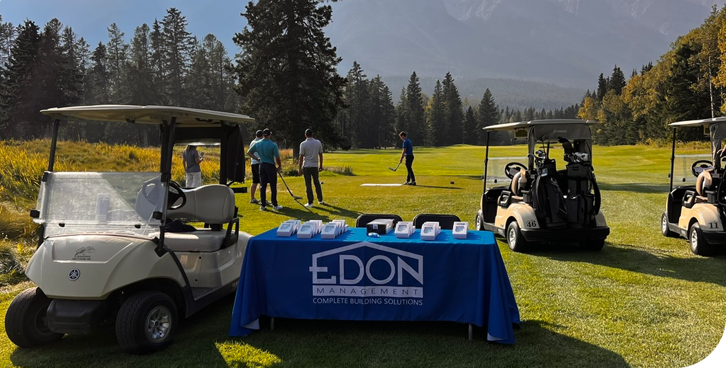 On a golf course, a table with EDON Management promotional materials is set up near golf carts. Golfers are practicing swings in the background surrounded by trees and mountains.