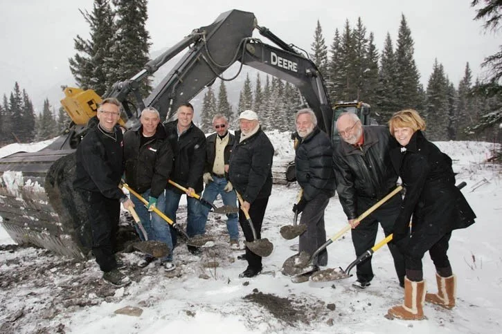 Group of eight people standing in the snow holding shovels in front of an excavator with