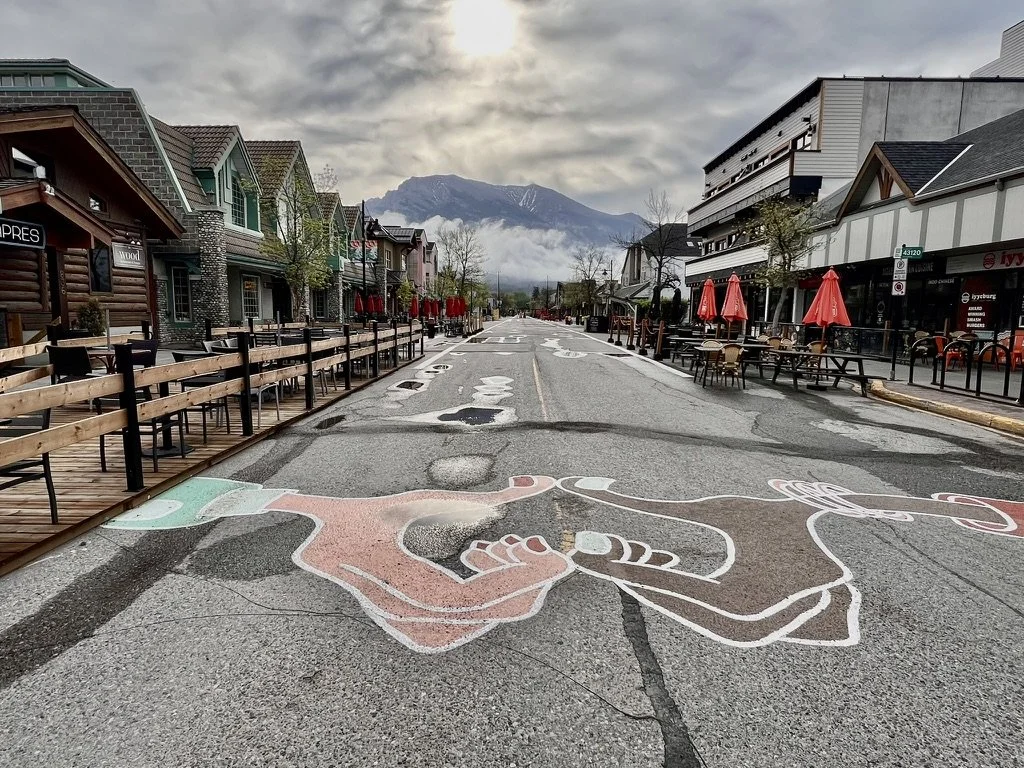 Downtown street with shops and outdoor seating, painted hood ornament design on the road, mountain in the background, overcast sky.