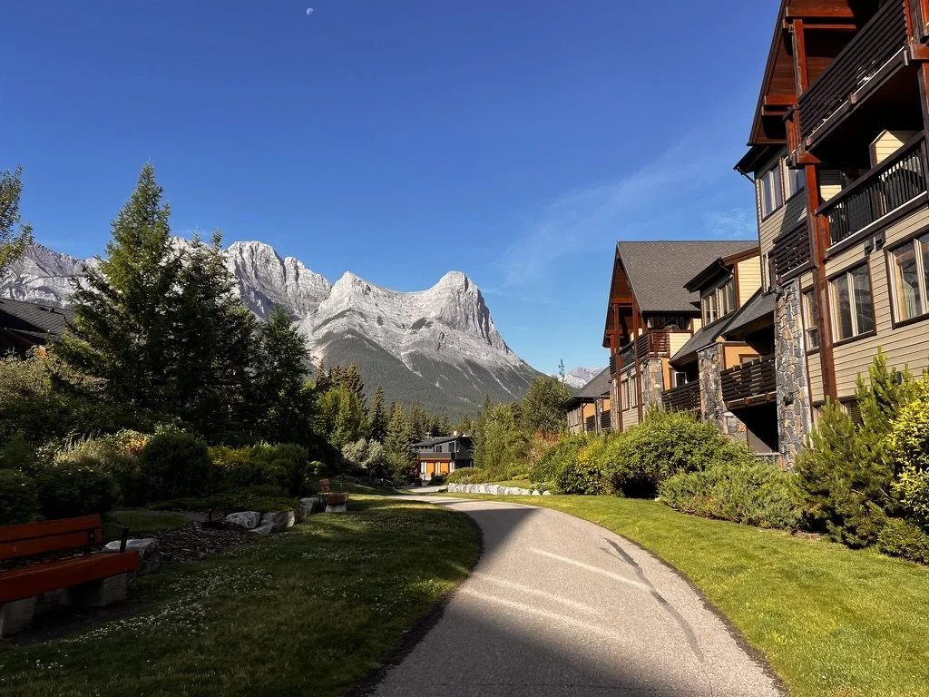 Residential buildings with balconies along a winding pathway, lush greenery, and mountains in the background under a blue sky.