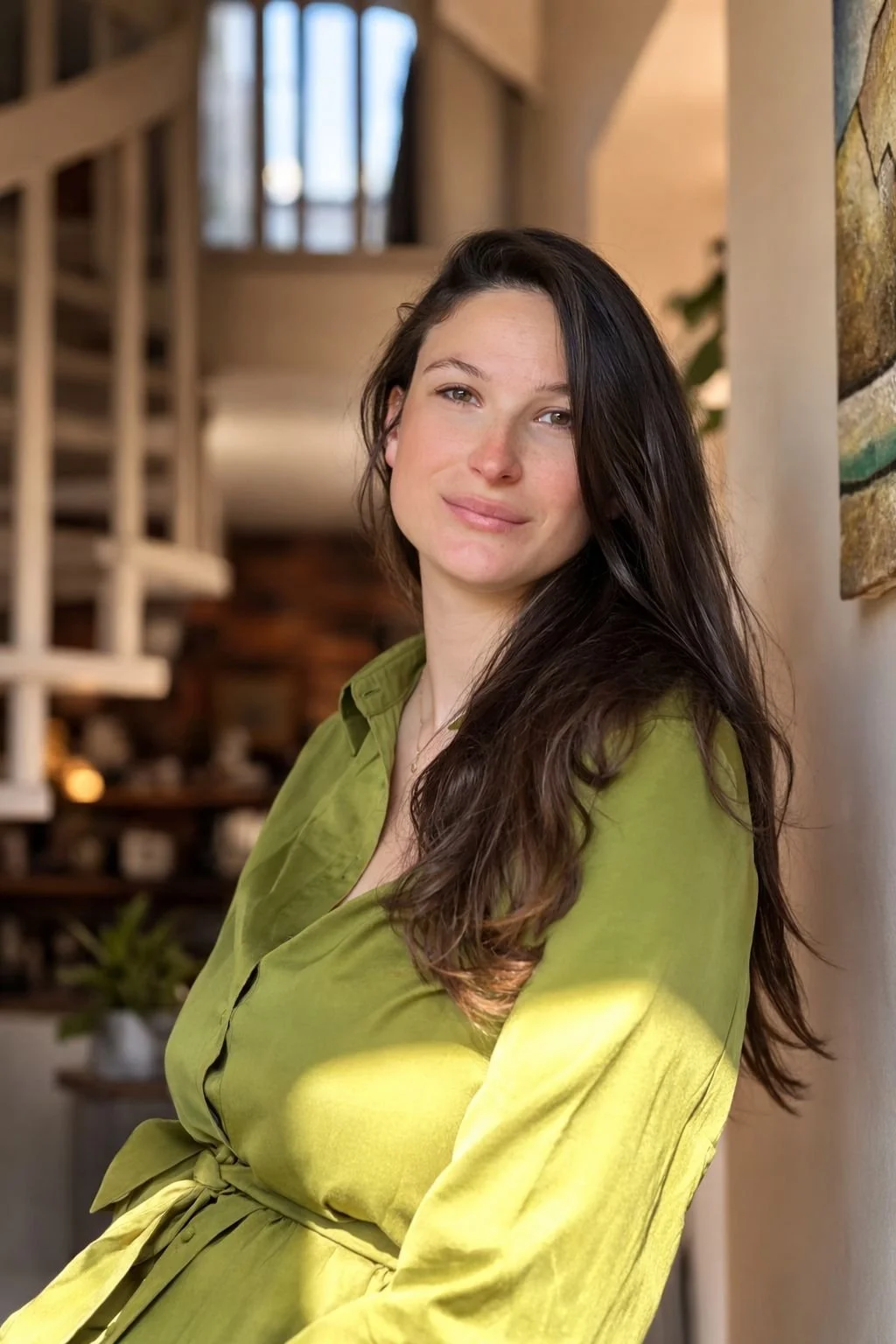 Jeune femme avec longs cheveux foncés et peau claire, portant une robe vert olive, posant dans une maison lumineuse.