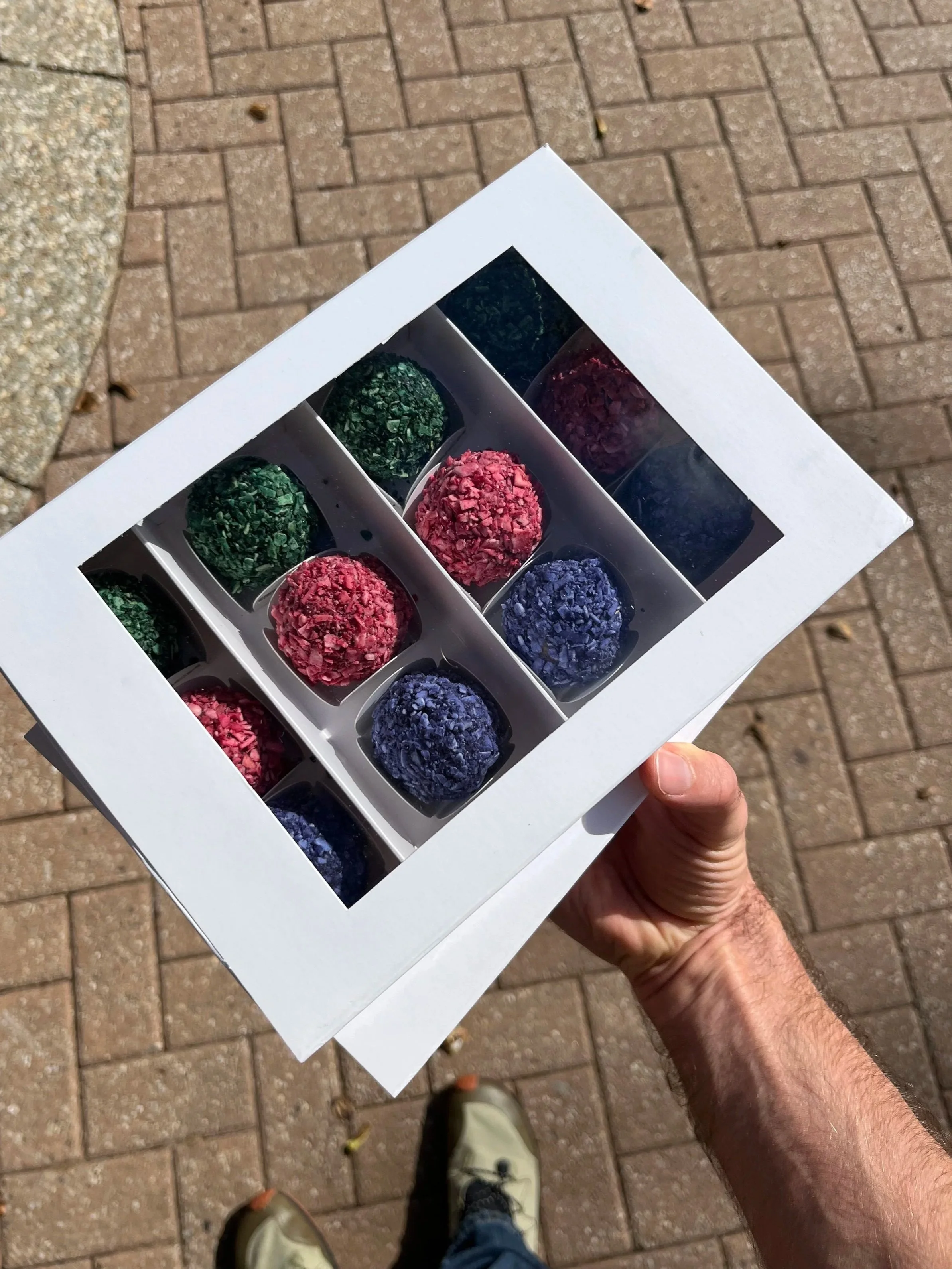 A person holding a white box of assorted colorful chocolates, viewed from above outdoors on a paved surface.