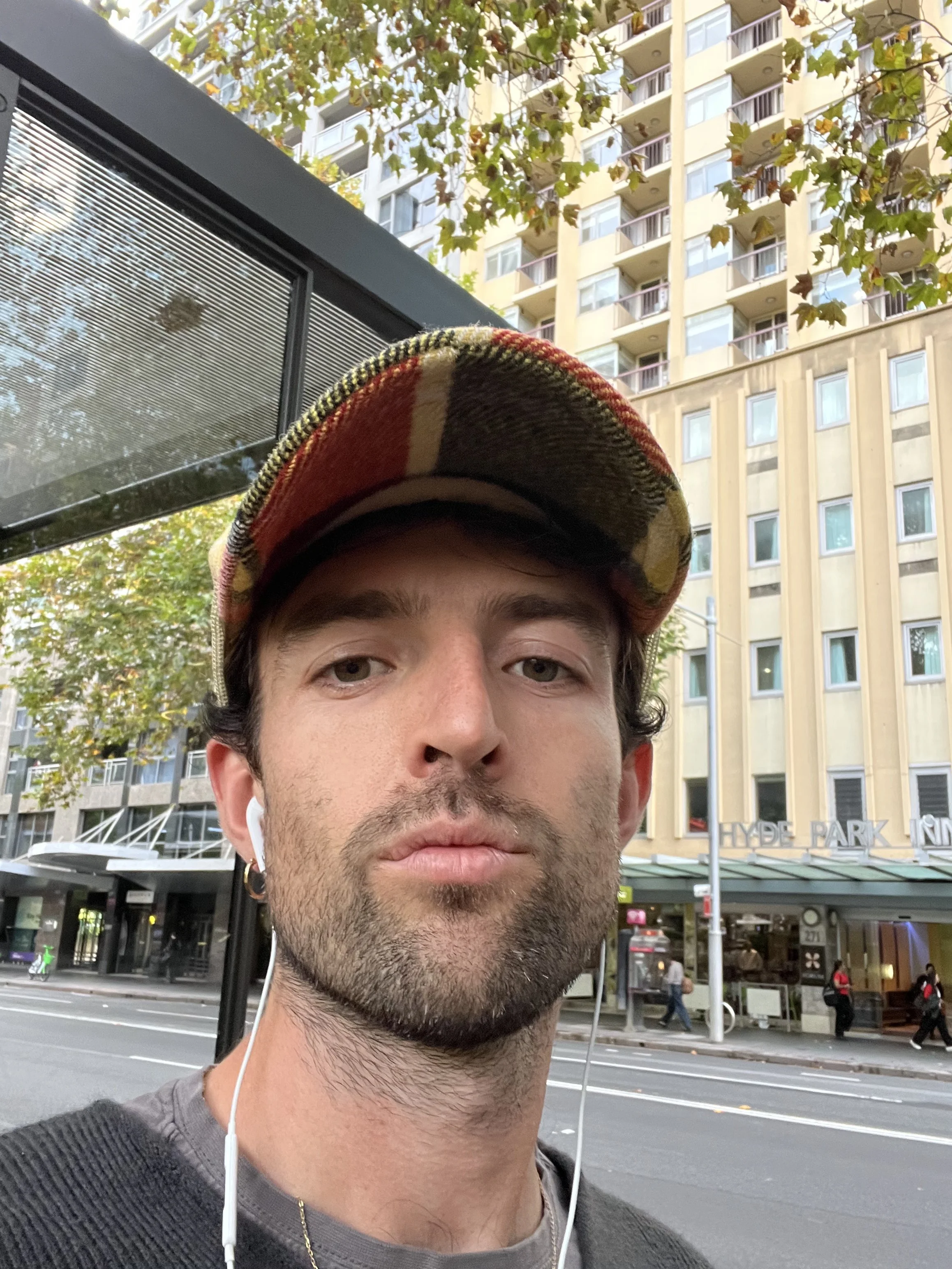 Close-up of a man wearing a plaid cap and white earbuds, with an urban city street and tall apartment buildings in the background.