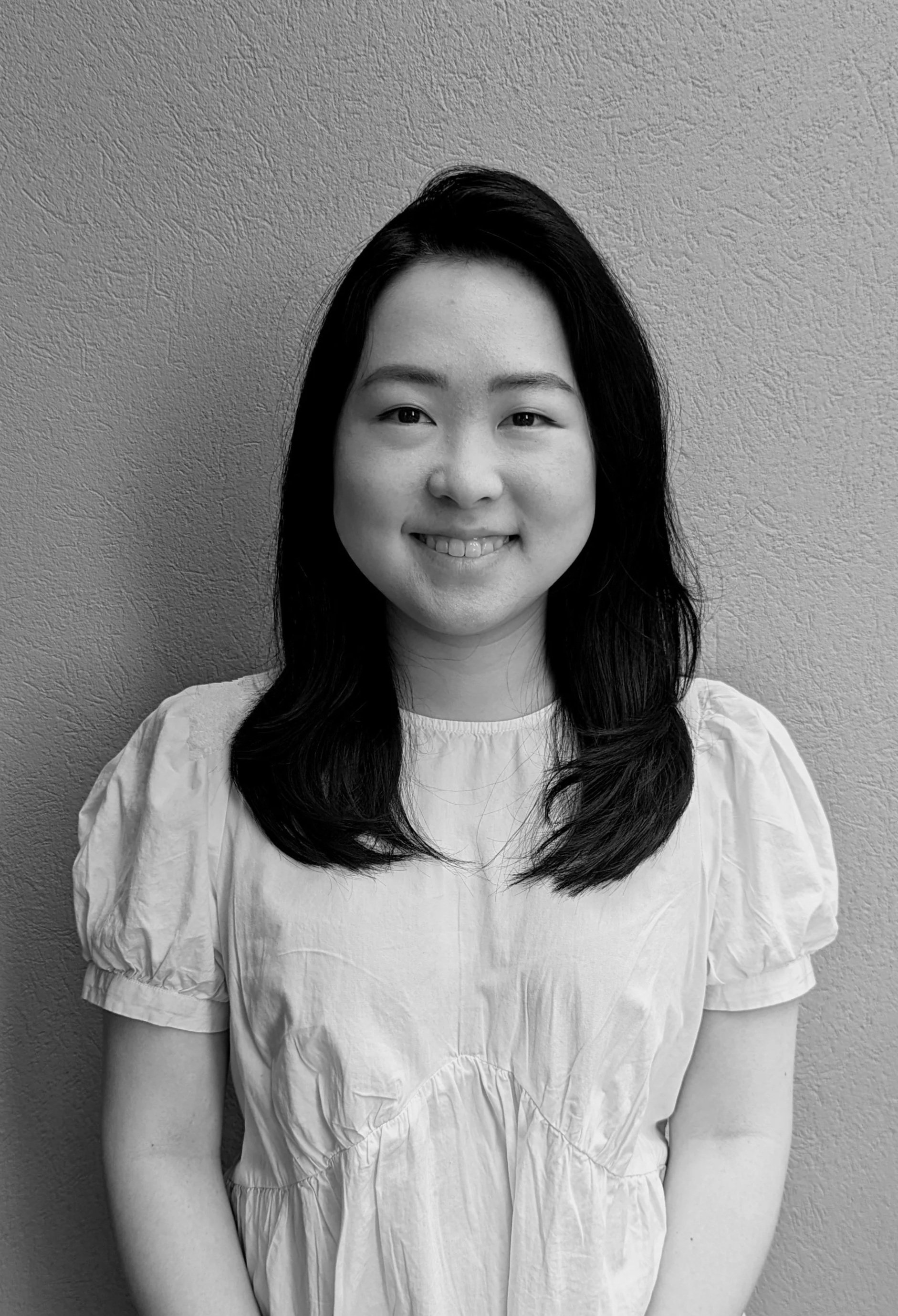 Black and white portrait of a young woman with long dark hair, smiling, wearing a light-colored top, standing against a textured wall.
