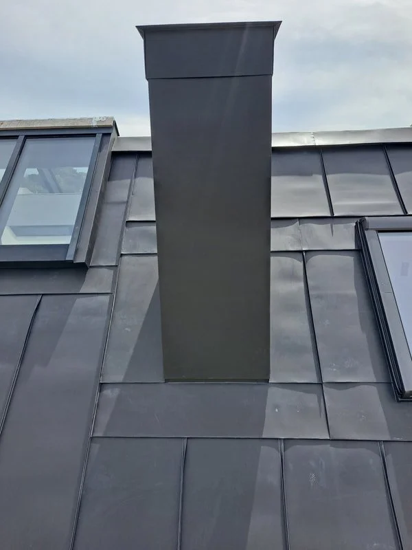 A close-up view of a modern roof with black metal tiles and a large chimney protruding from the roof. The sky is cloudy.
