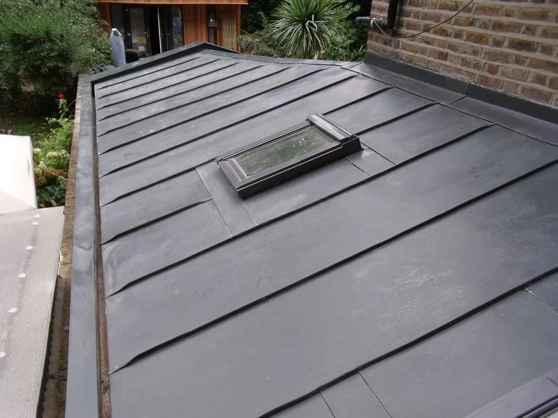 Metal roof with a skylight window surrounded by green plants and brick wall