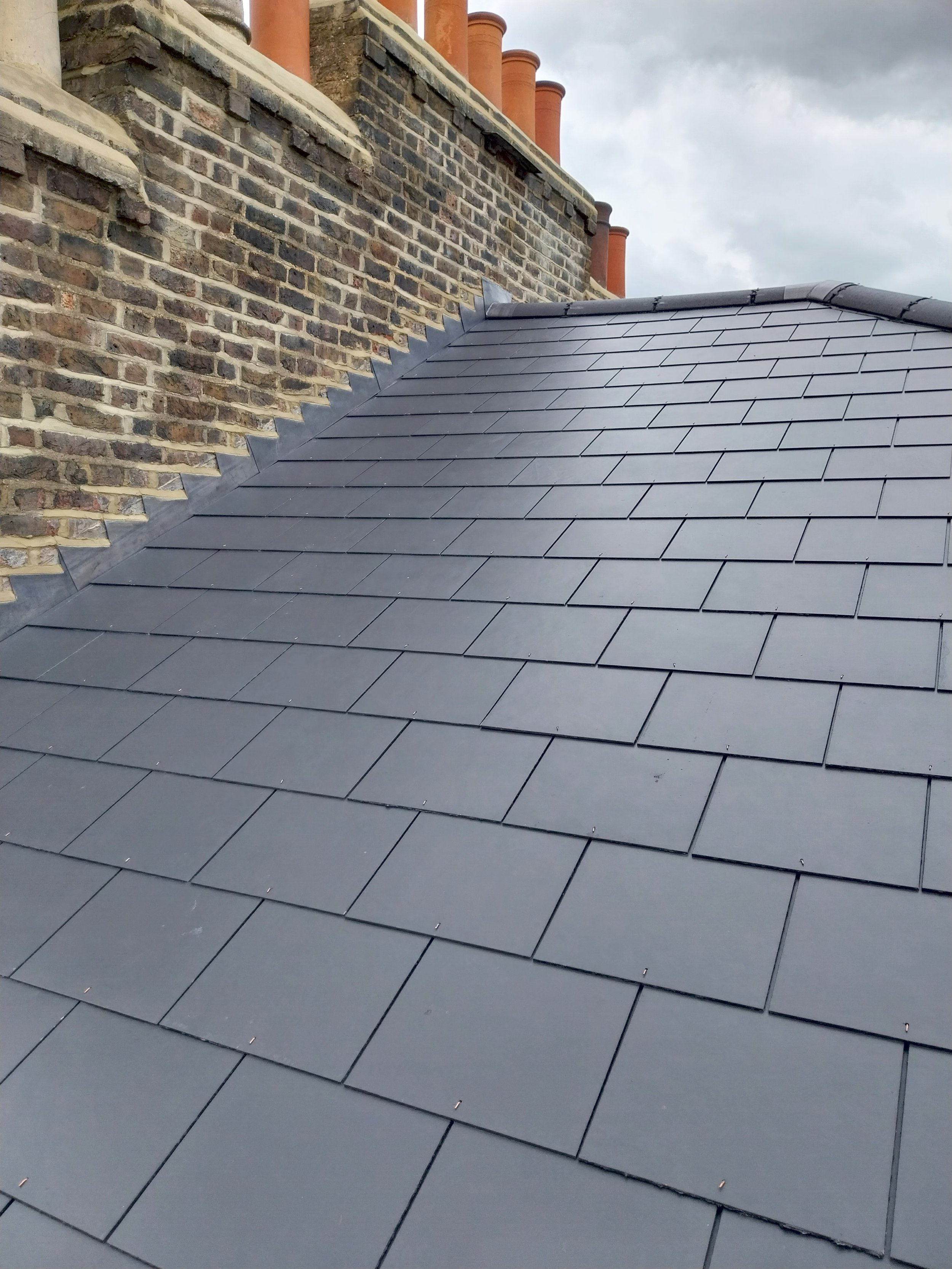 Close-up of a roof with slate tiles, a brick chimney wall with multiple red clay chimney pots.
