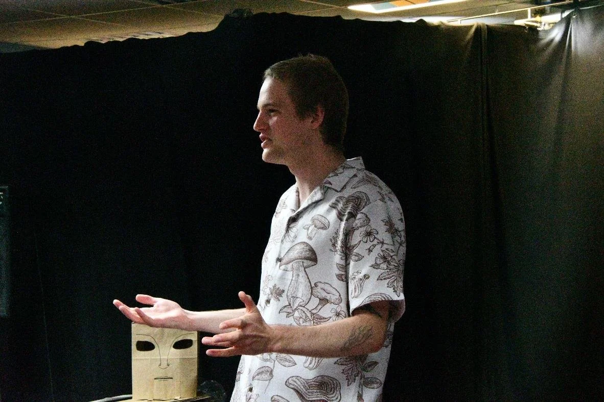 A young man in a white shirt with mushroom and nature prints, speaking or presenting with hands gesturing, standing in front of a black background with a wooden mask on a stand.