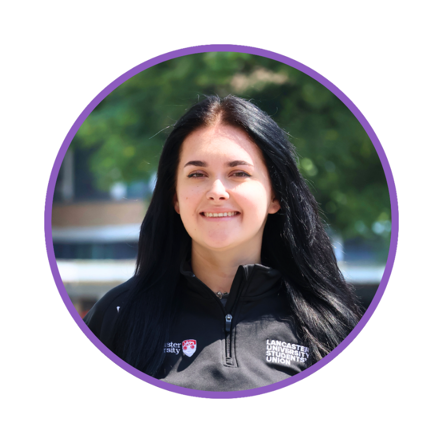 Young woman with long black hair smiling outdoors in front of green trees, wearing a Lancaster University Students' Union jacket.