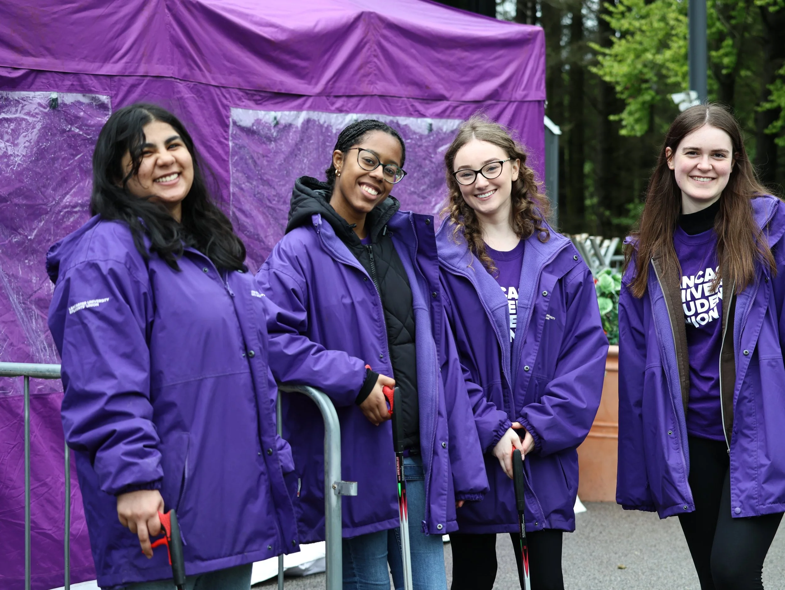 Four young women wearing purple jackets and standing outdoors in front of a purple tent, smiling.