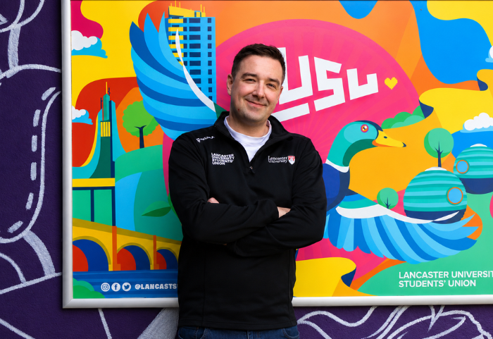 A young man standing in front of a colorful mural at Lancaster University. The mural features abstract designs, a duck, a building, trees, and a pink background with white text. The man is smiling, wearing a black jacket with a university logo, and has his arms crossed.