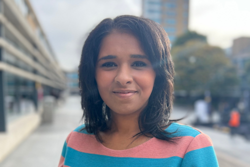 Young woman with shoulder-length dark hair, smiling, wearing a striped pink and blue shirt, standing outdoors on a city street with buildings and trees in the background.