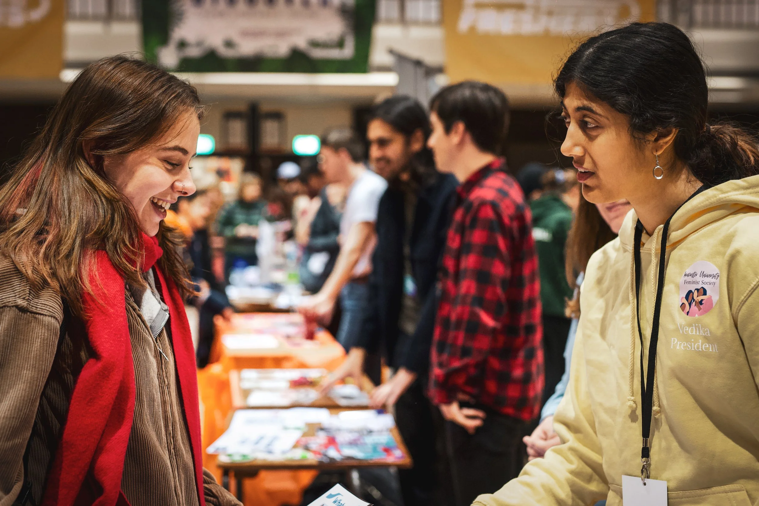 Two women engaging in conversation at an indoor event, with people and tables with informational materials in the background.
