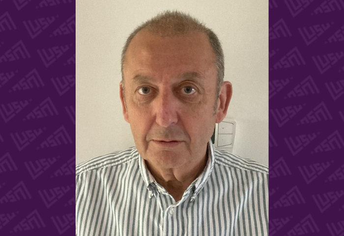A middle-aged man with short, balding hair wearing a vertically striped button-up shirt, looking directly at the camera in front of a plain background.