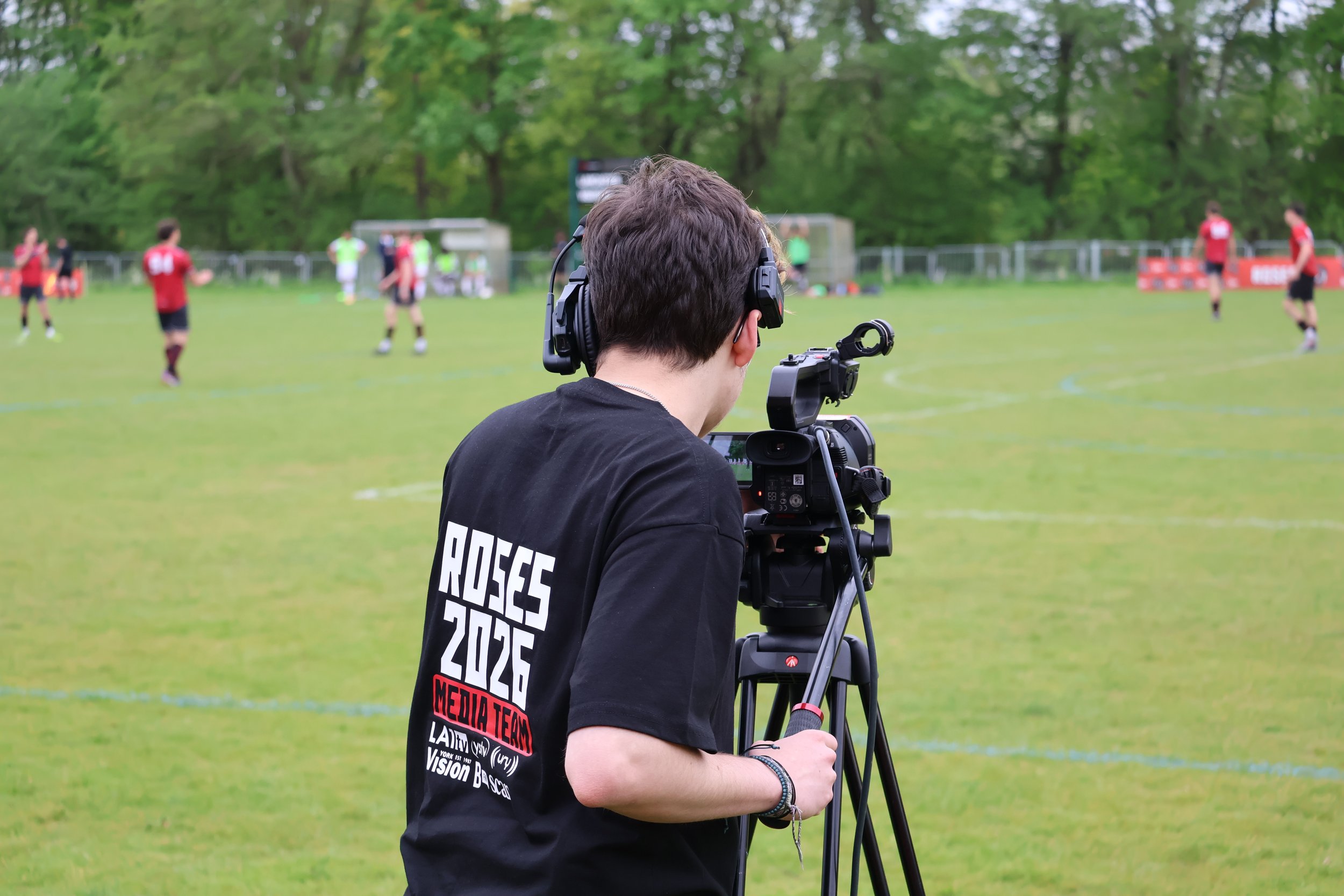 A person with headphones operating a professional video camera on a tripod, filming a sports game on a grass field with players in red jerseys.