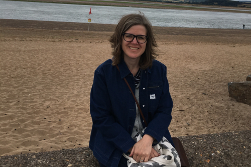 A woman with glasses and shoulder-length hair smiling on a sandy beach near a body of water.