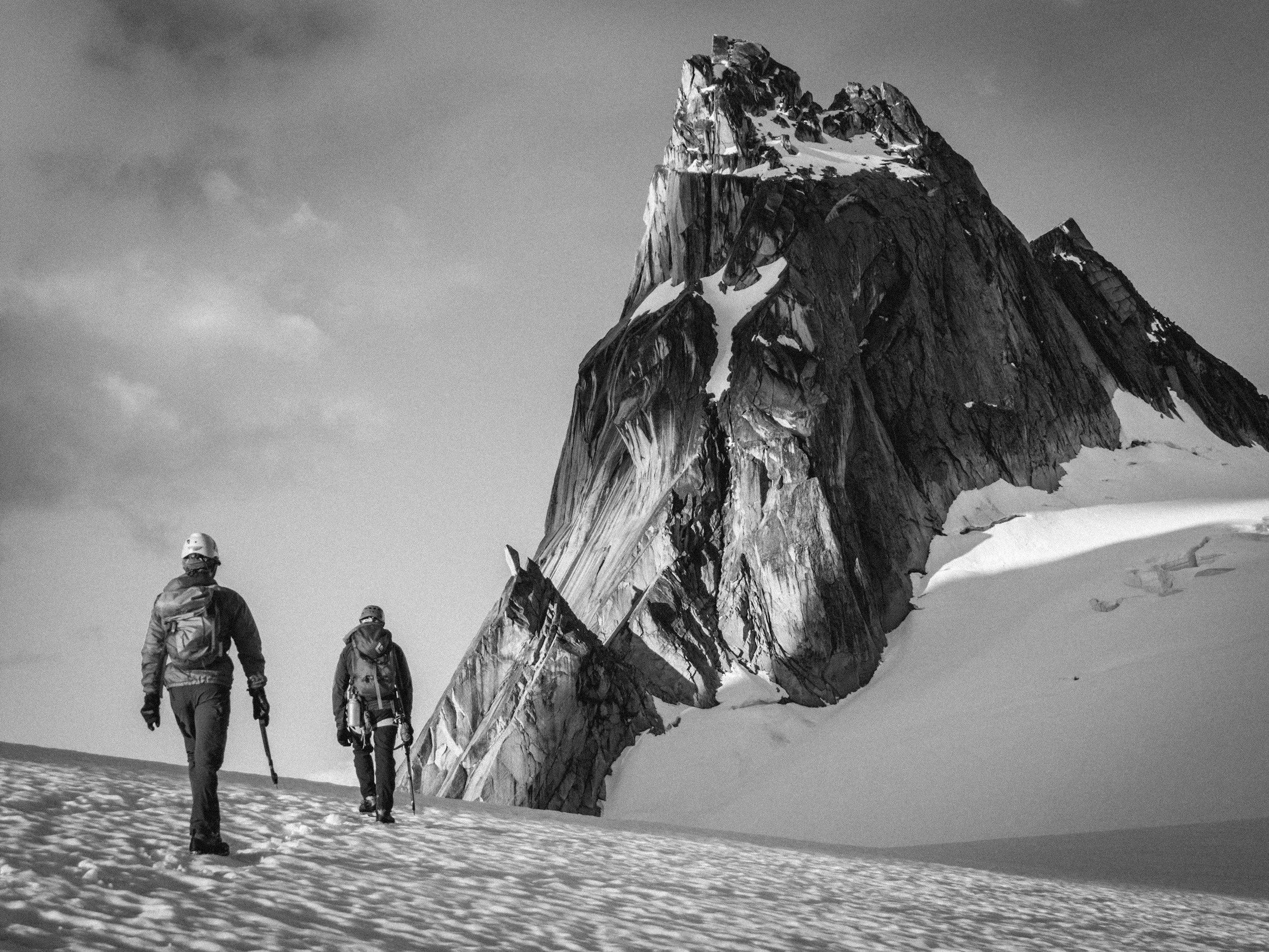 Black and white image of two climbers in full gear approaching the base of a steep, lightly snow-covered mountain.