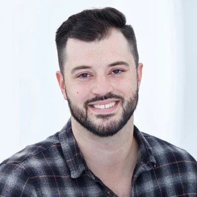 Head and shoulders portrait of Iain Alexander, Director of IGA Consulting. A white male in his mid-30s, he has short, dark brown hair and a full, close-cropped beard. He is wearing a lumberjack check shirt in black, orange, and grey.