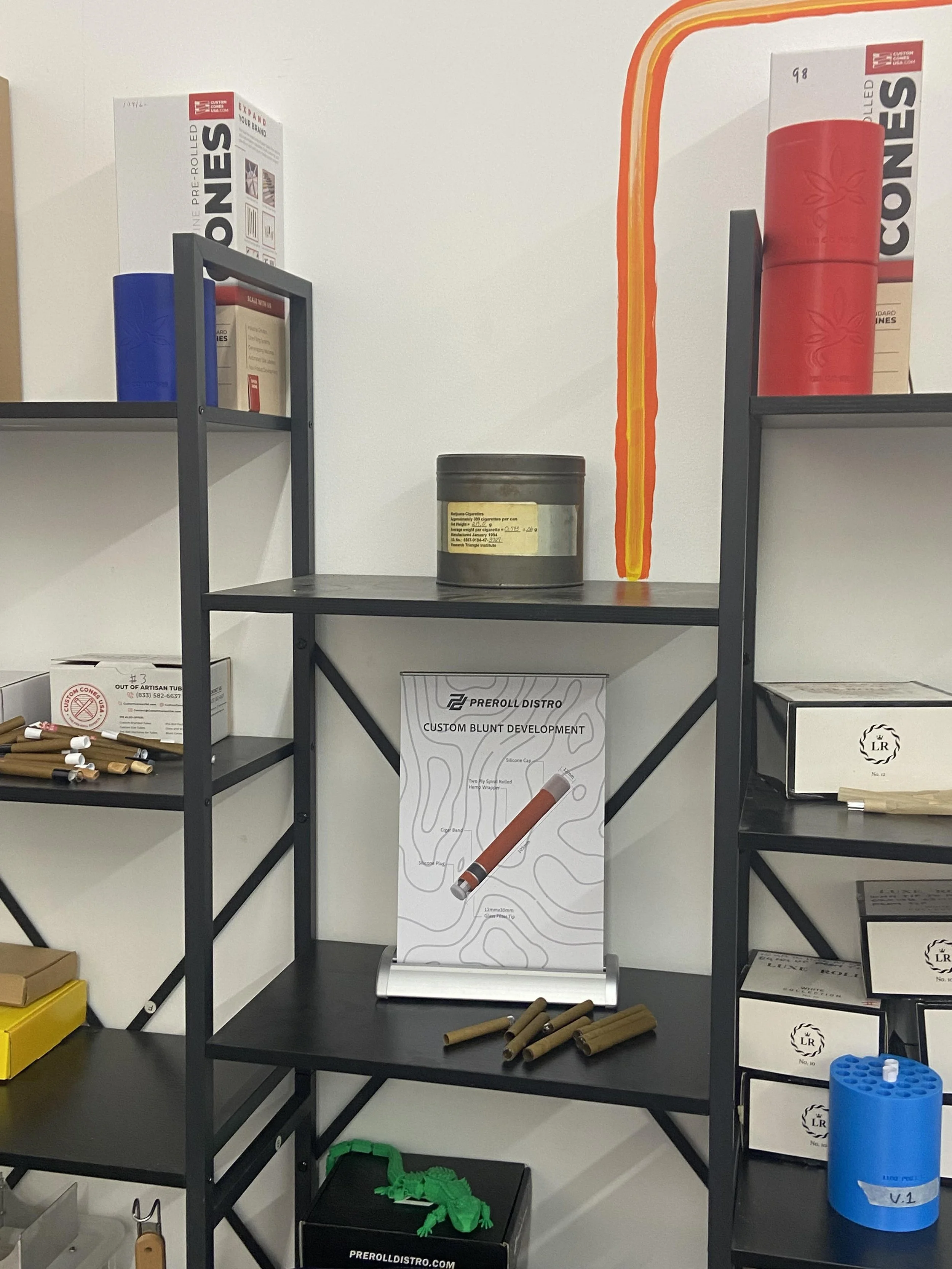 A metal cannabis tin displayed on a shelf among cone packaging and Hummingbird tools.