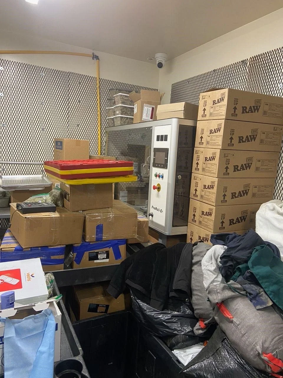 Preroll equipment surrounded by stacked cone boxes, supplies, and bags in a storage area.