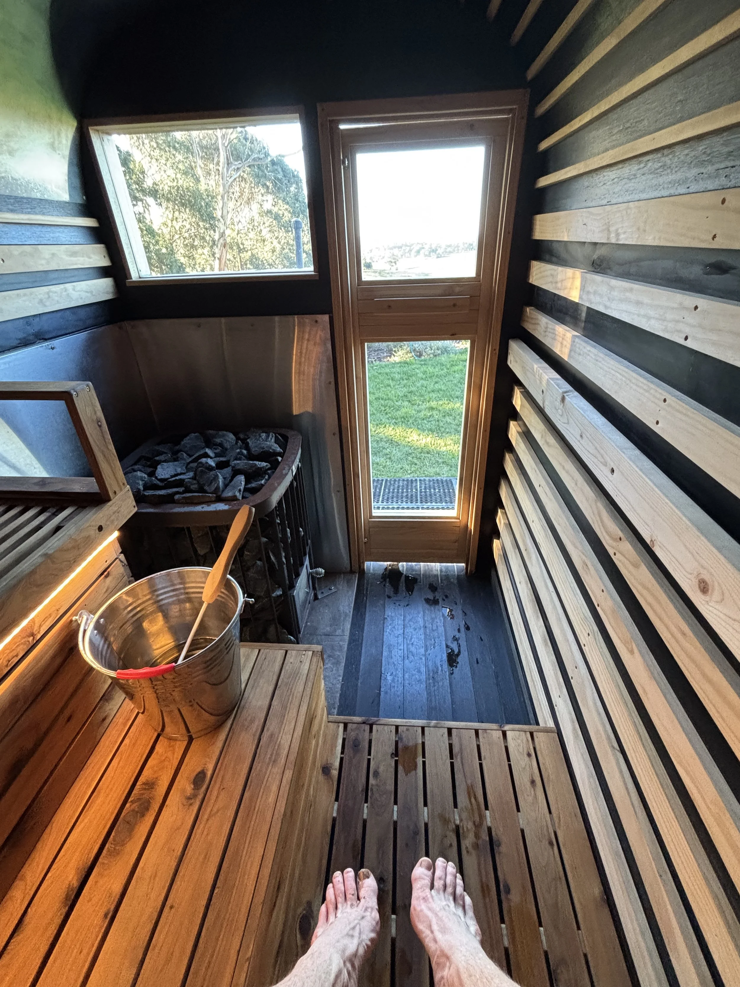 View from inside a wooden mobile sauna with a person's feet visible at the bottom, a bucket with a ladle, and two windows showing trees and grass outside, a pleasant view.
