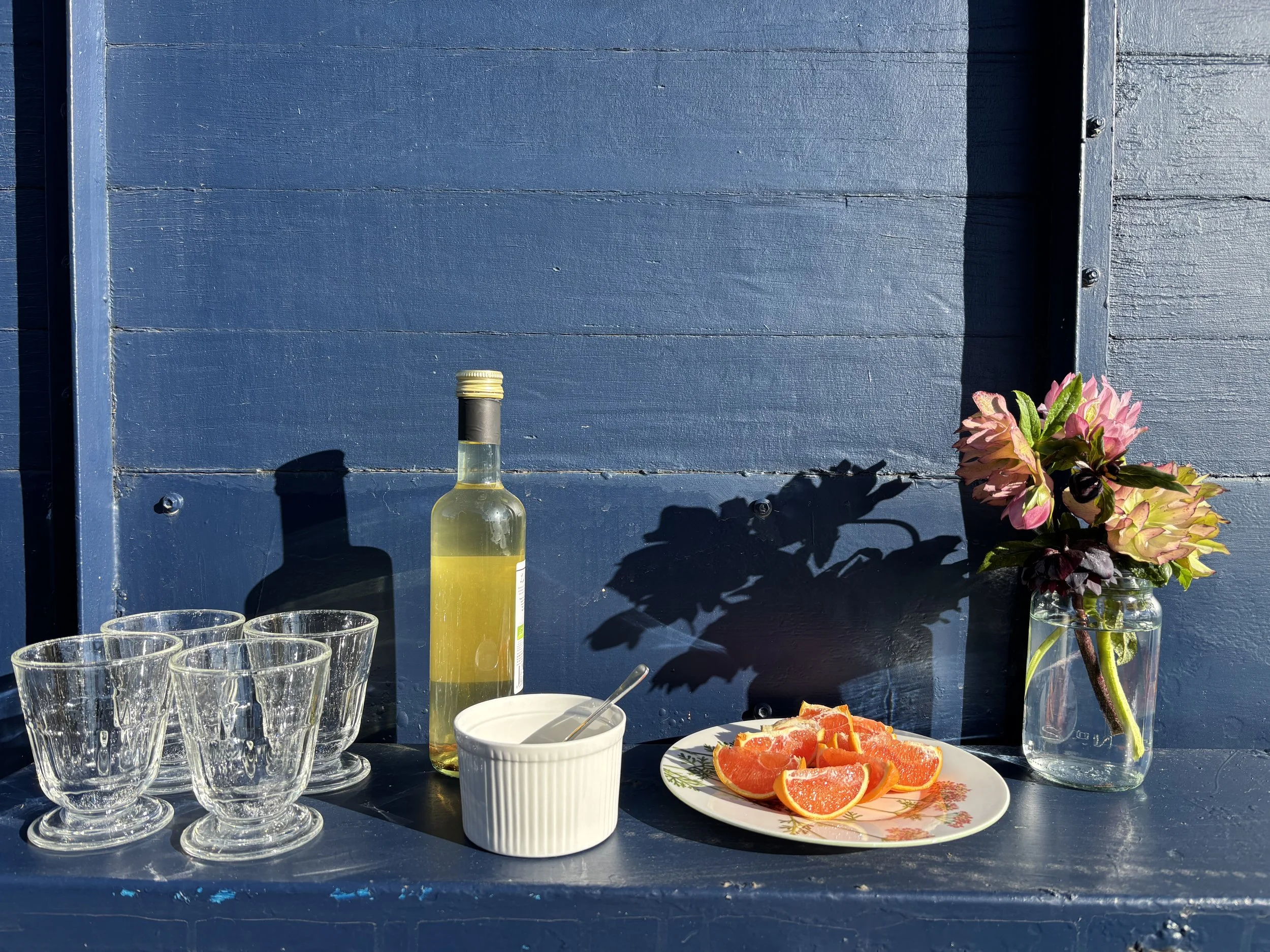 A shelf on the outside of the mobile sauna's blue wooden wall, with a group of empty clear glasses, a bottle of apple cider vinegar wine, a white bowl with a spoon, a plate of sliced oranges, and a vase with pink flowers.