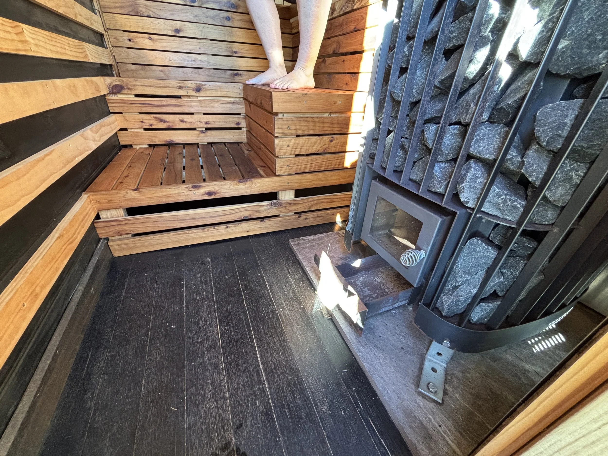 Feet of a person sitting on a wooden platform inside a mobile sauna, with wooden walls and a wood fired stone heater on the right side, with sunlight streaming through the sauna windows.
