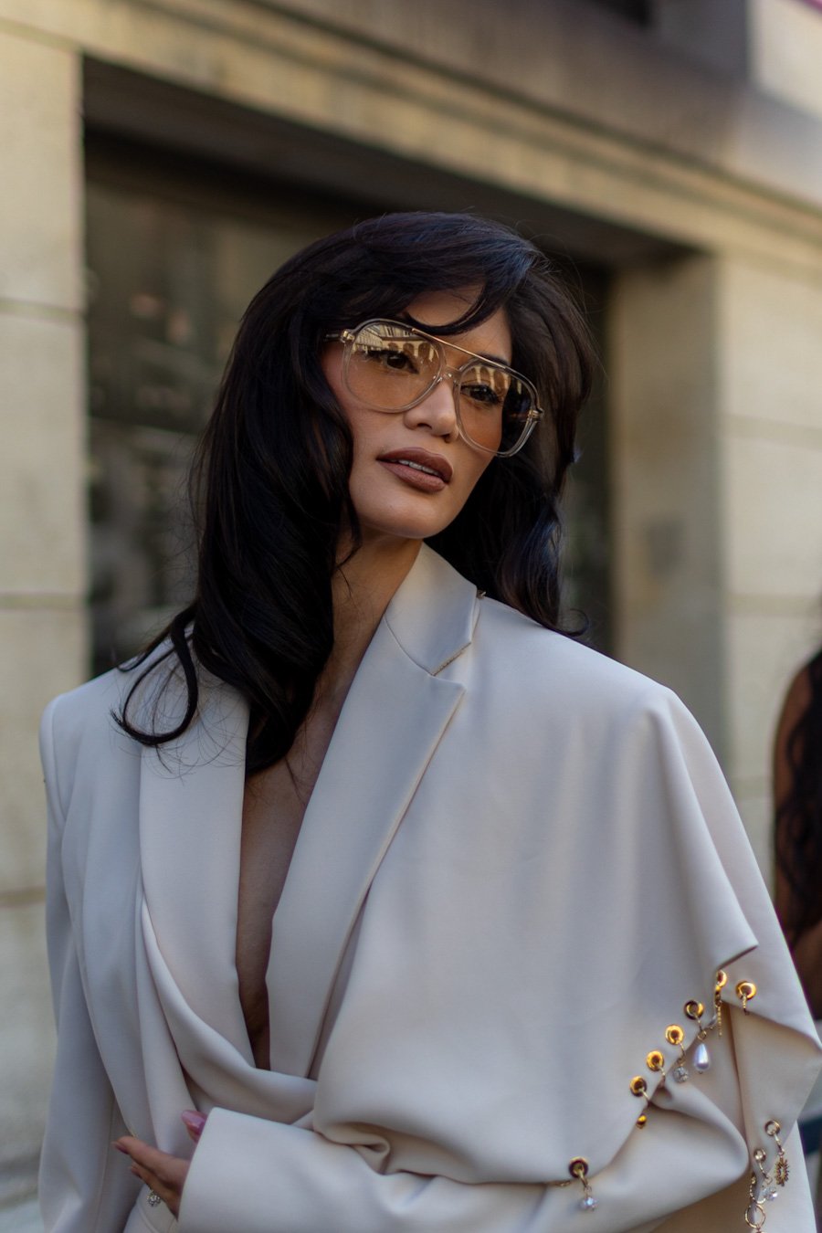 A woman with black hair wearing large aviator sunglasses and a cream-colored blazer with decorative embellishments on the sleeve, standing outside in front of a stone building.