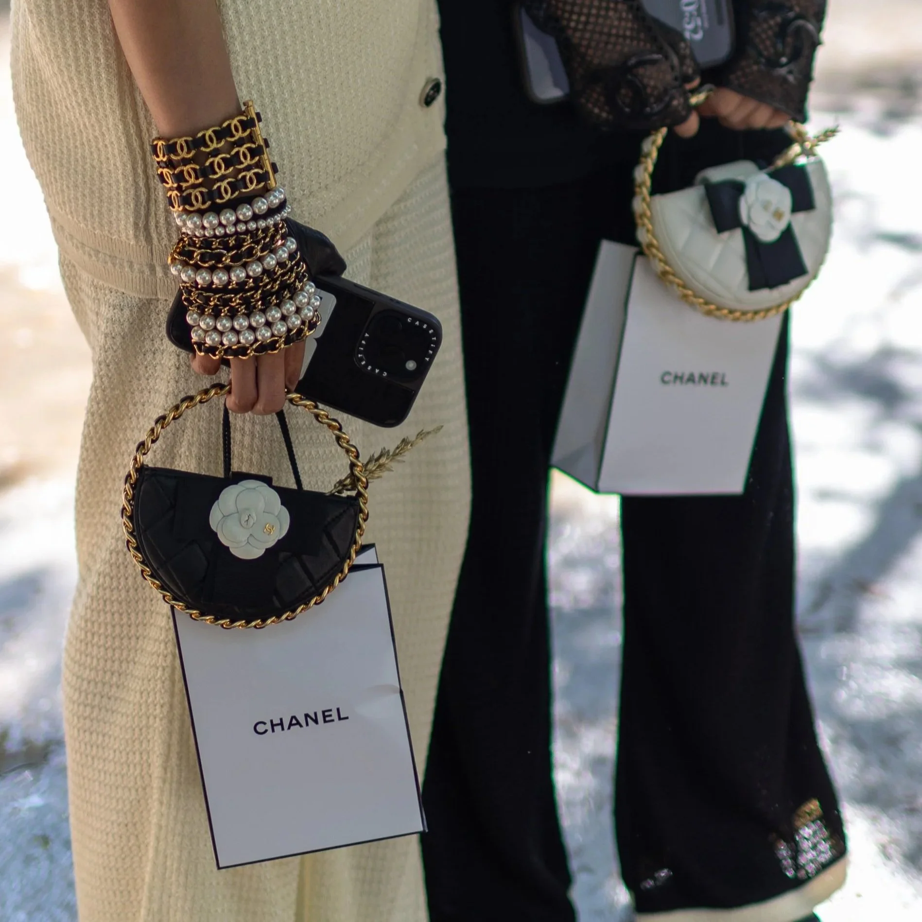 Close-up of two people holding Chanel branded shopping bags and handbags with decorative jewelry, including pearl and chain bracelets, in an outdoor setting.