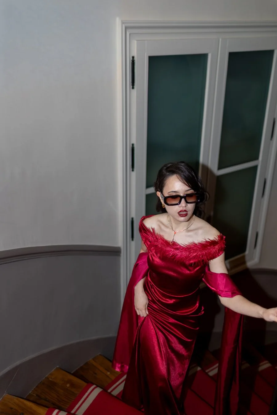 Woman in a red satin dress with feather trim on the neckline, wearing dark sunglasses, walking down a staircase next to a white wall and glass-paneled door.