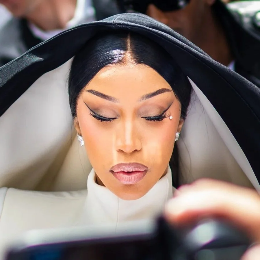 Close-up of a woman with dark hair, styled with a side part, wearing makeup with winged eyeliner and natural-colored lipstick, looking down at a phone or device she is holding.